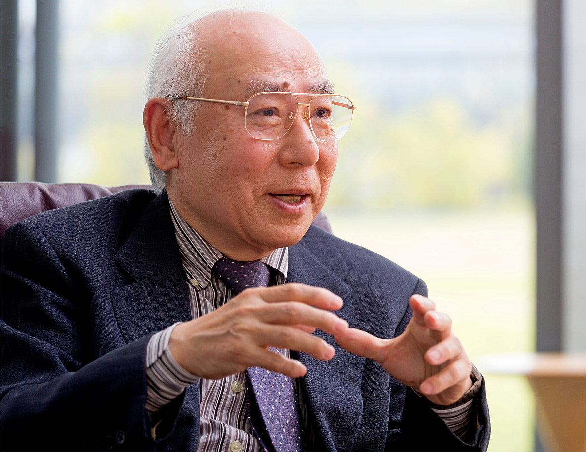 portrait of an elderly Japanese man wearing a blue suit, a striped shirt, and polka-dotted tie. He has gold-rimmed glasses, and looks like he's in the middle of explaining something.