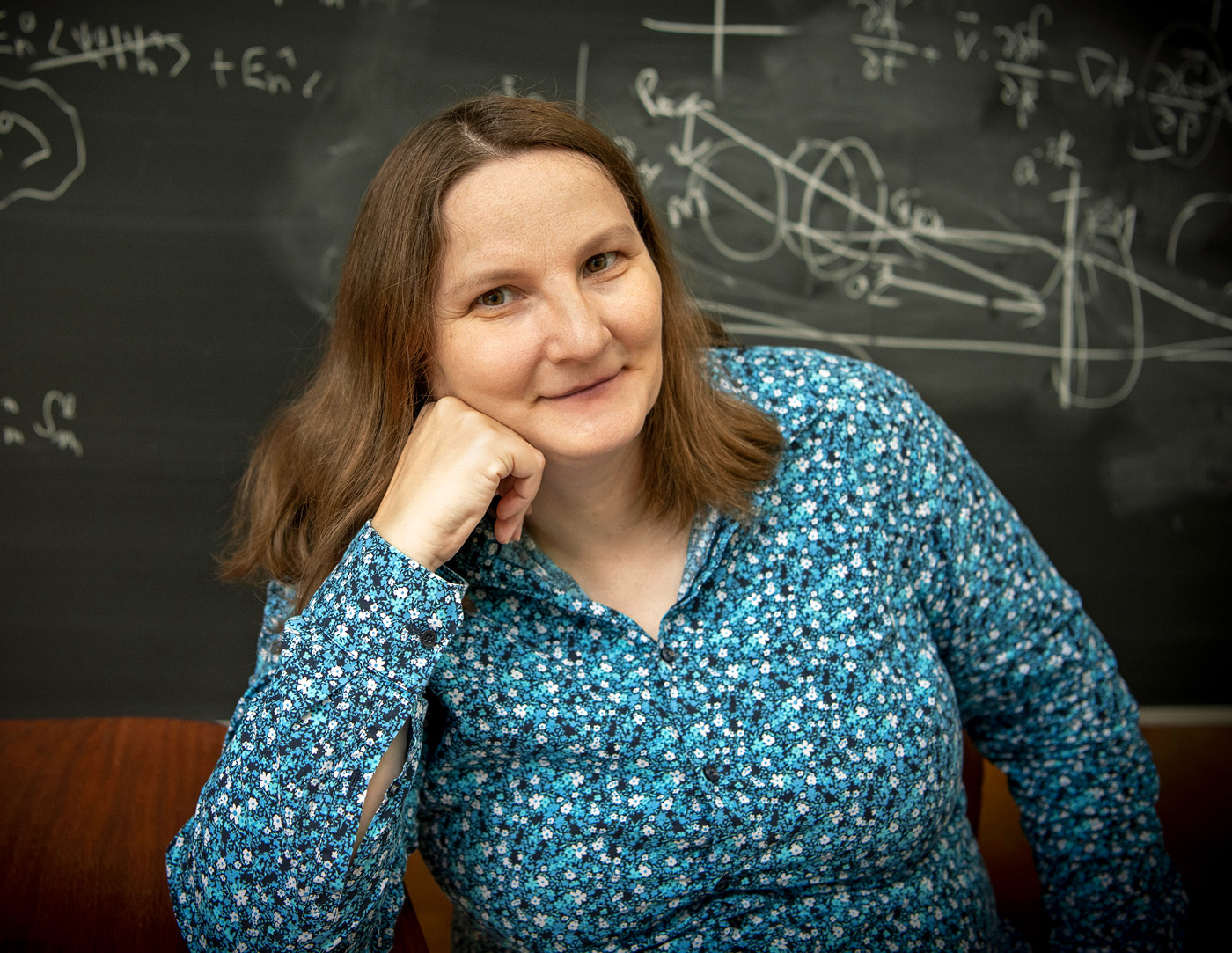 Photo of a woman in a floral blouse sitting in front of a blackboard.