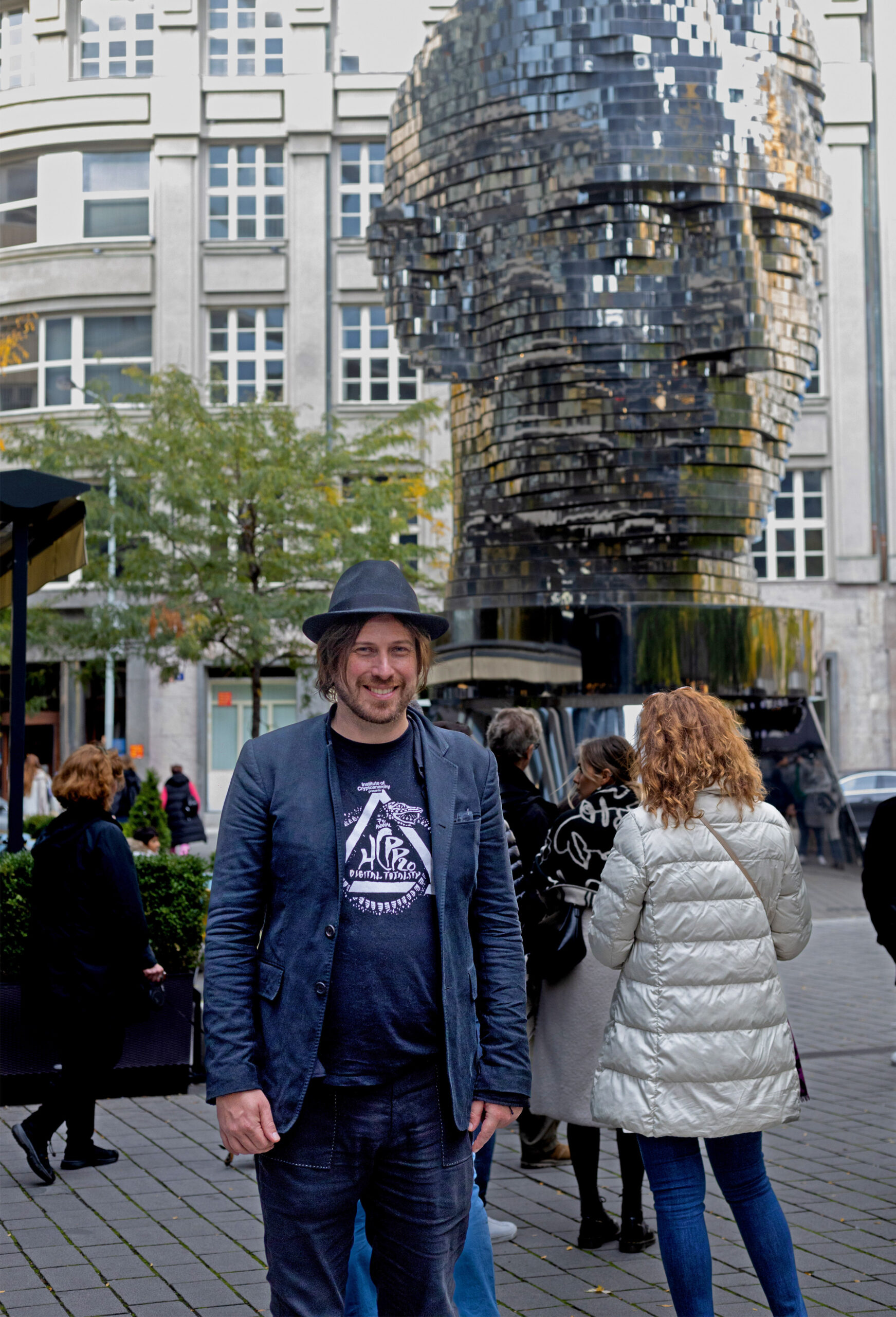 Harry Halpin in T-shirt blazer and Trilby in front of a metal statue in a public square