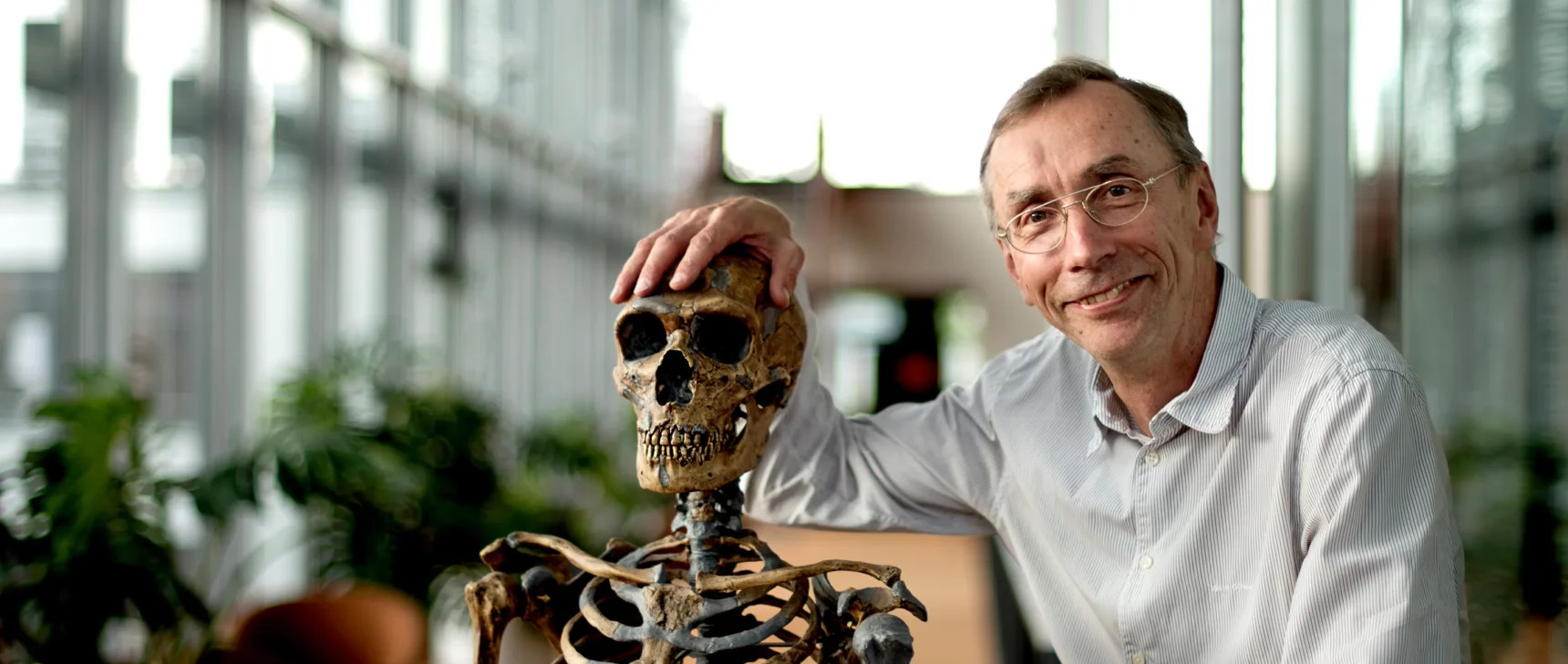 Photo of man with gold-rimmed glasses and a pin-striped button-up shirt posing with his hand on an early hominid skeleton