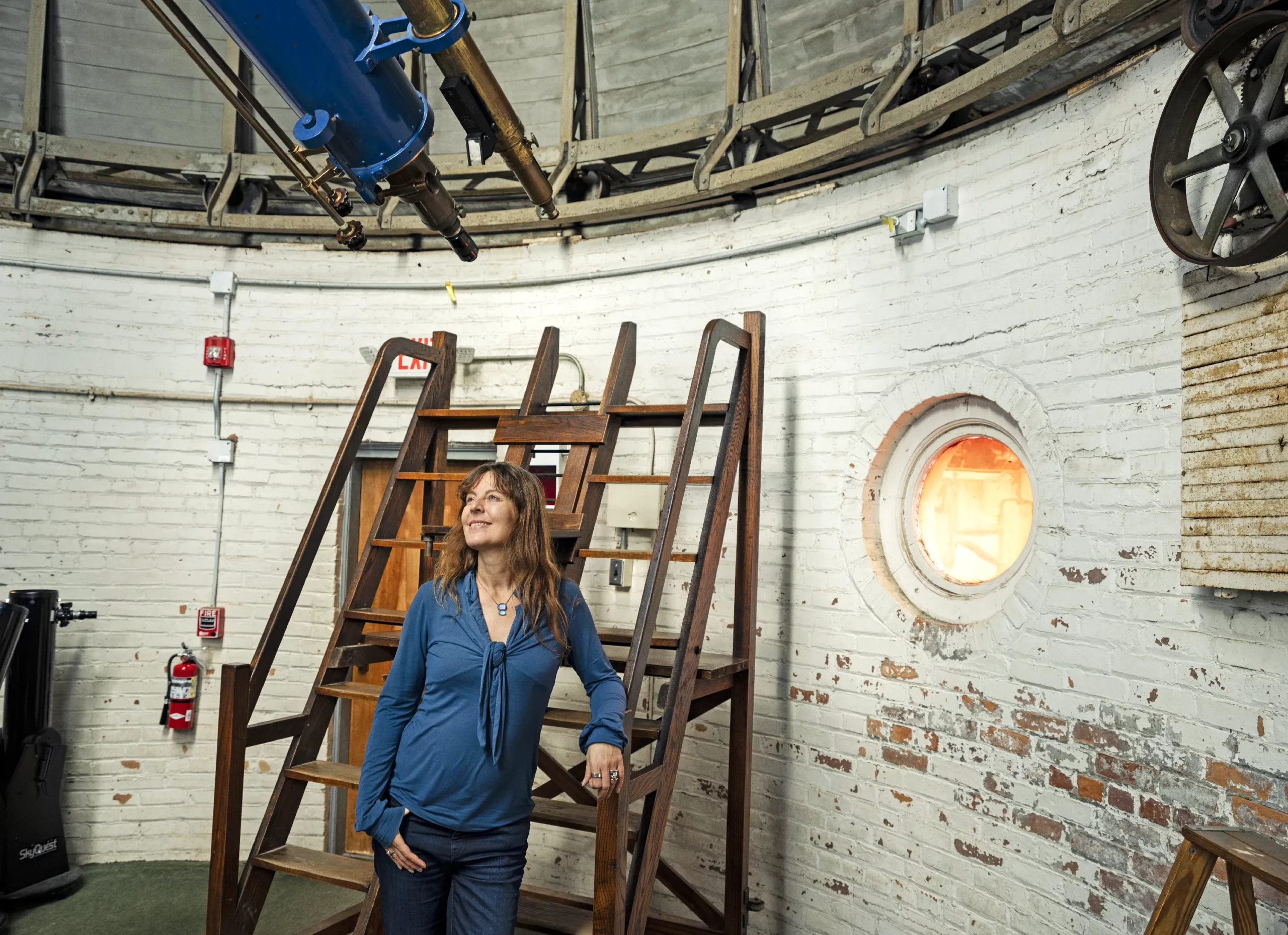 Kaltenegger leans against wooden stairs in the round, brick-walled room of an observatory. Part of a telescope appears at the top of the photo.