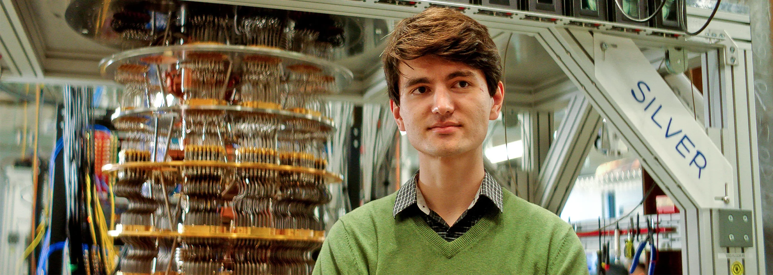 A young man with brown hair in a green sweater sits in front of a cryostat.