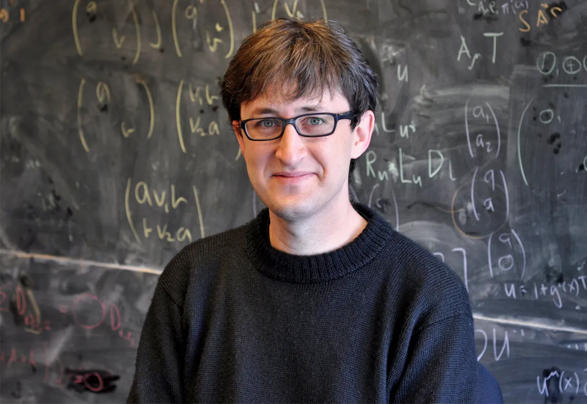 A man in glasses and a black sweater stands in front of a blackboard and smiles at the camera.