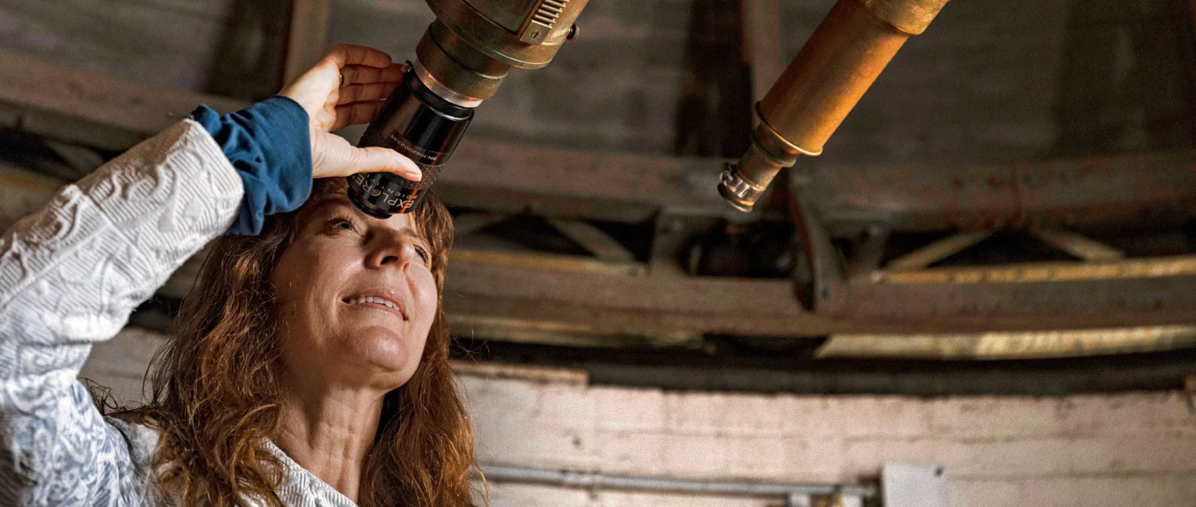 Lisa Kaltenegger, a woman with red hair, peers through the eyepiece of an antique telescope.