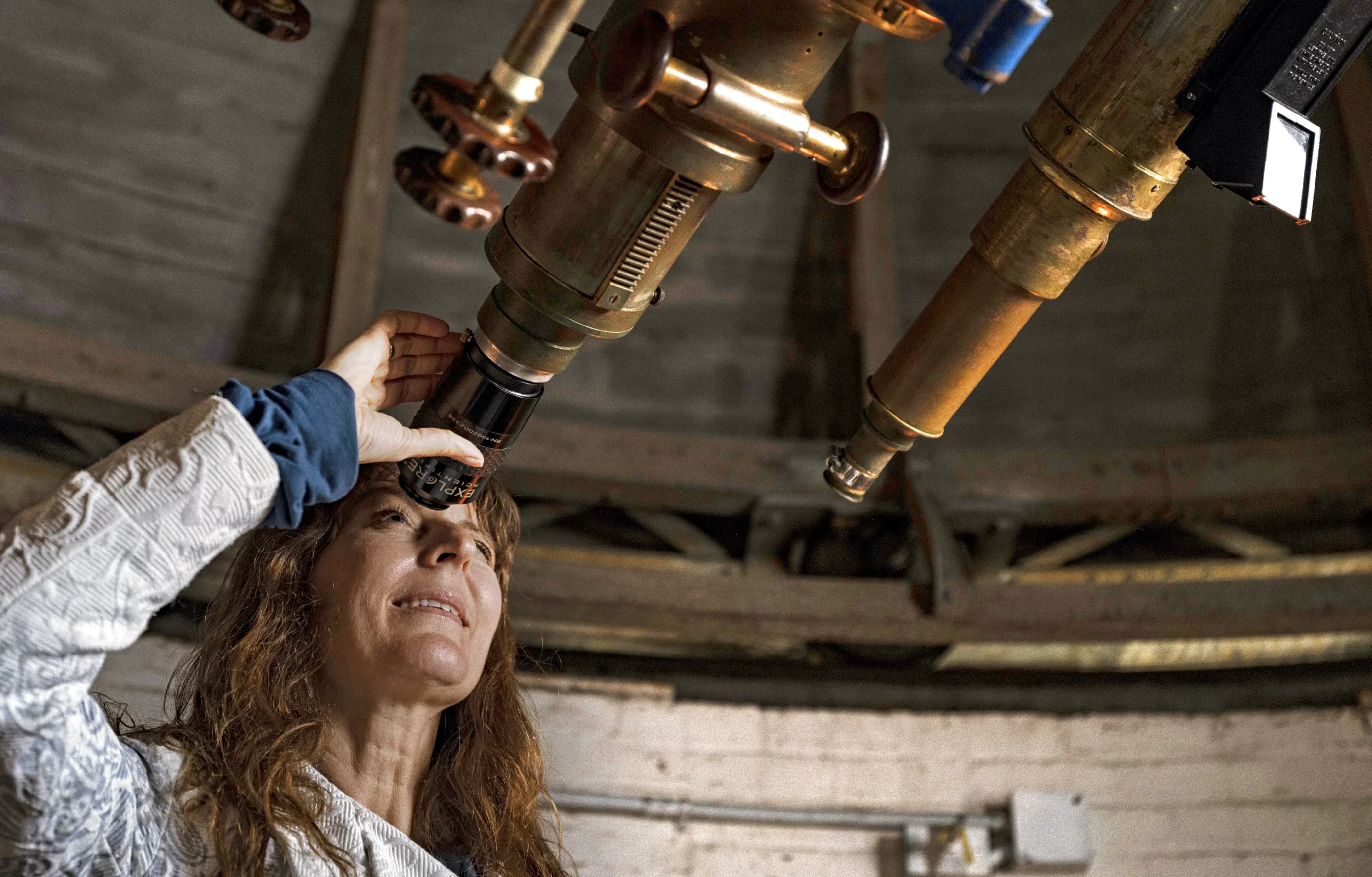 Lisa Kaltenegger, a woman with red hair, peers through the eyepiece of an antique telescope.