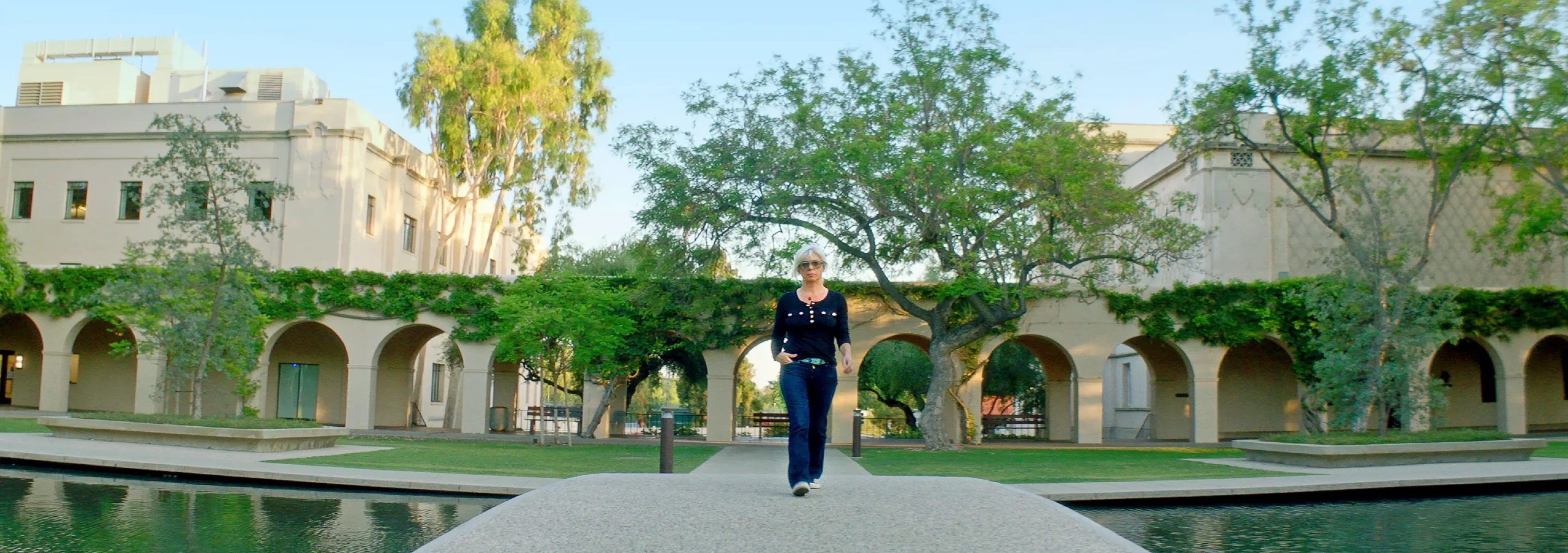 A woman walks down a path near some buildings and a reflecting pool.