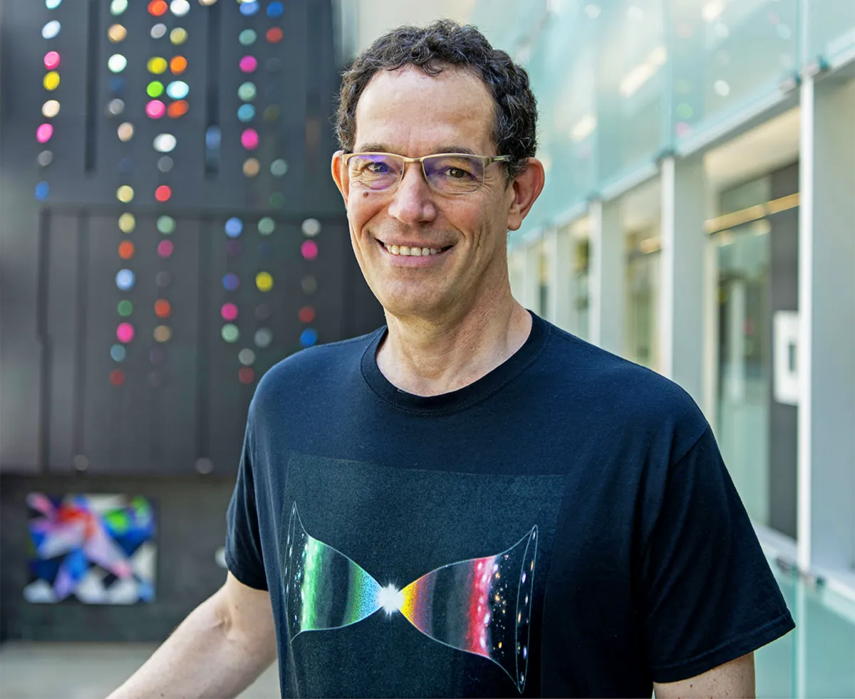 : A man with short curly hair smiles at the camera. His t-shirt displays a universe expanding outward from a Big Bang in two directions, to represent his mirror universe model.