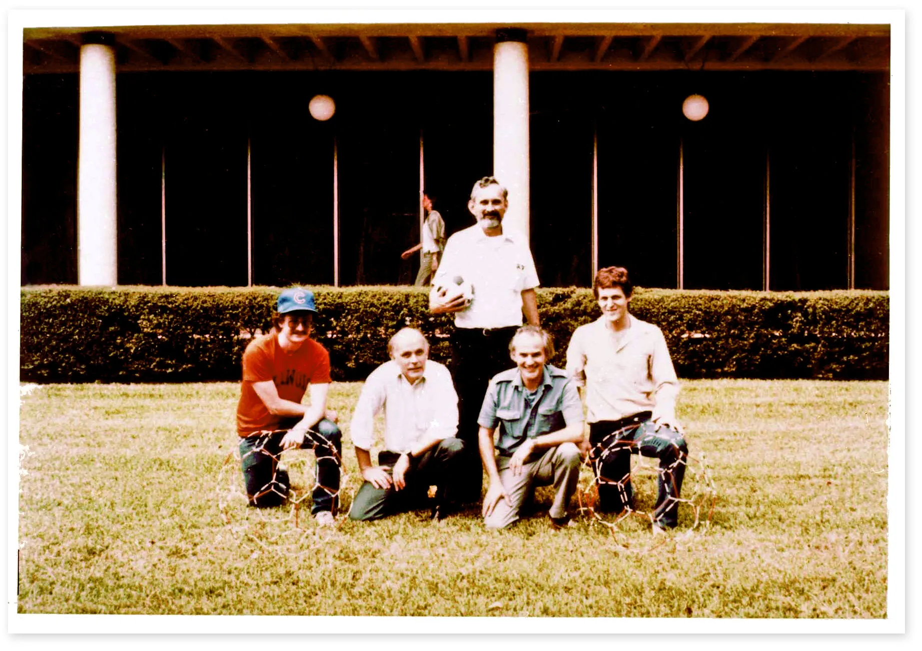 A 1980s-era photograph of four men kneeling and one man standing on the grass in front of a building. A ball-and-stick model of a buckyball rests on the grass.