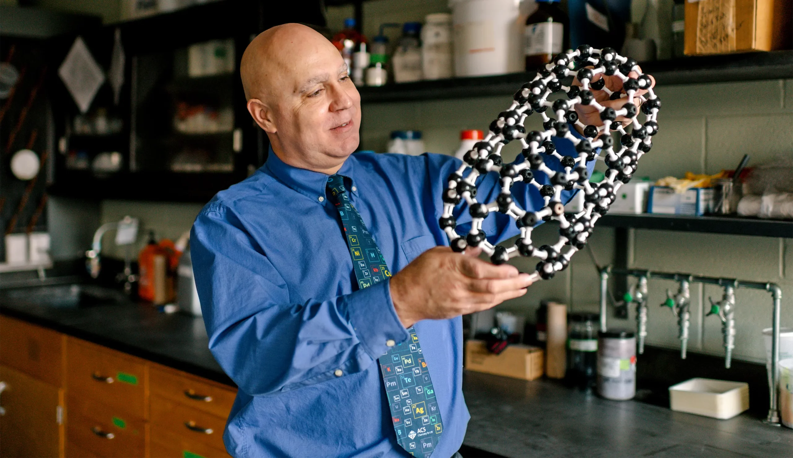 A bald man in a blue shirt and a periodic table tie holds up a pill-shaped ball-and-stick model of a fullertube.