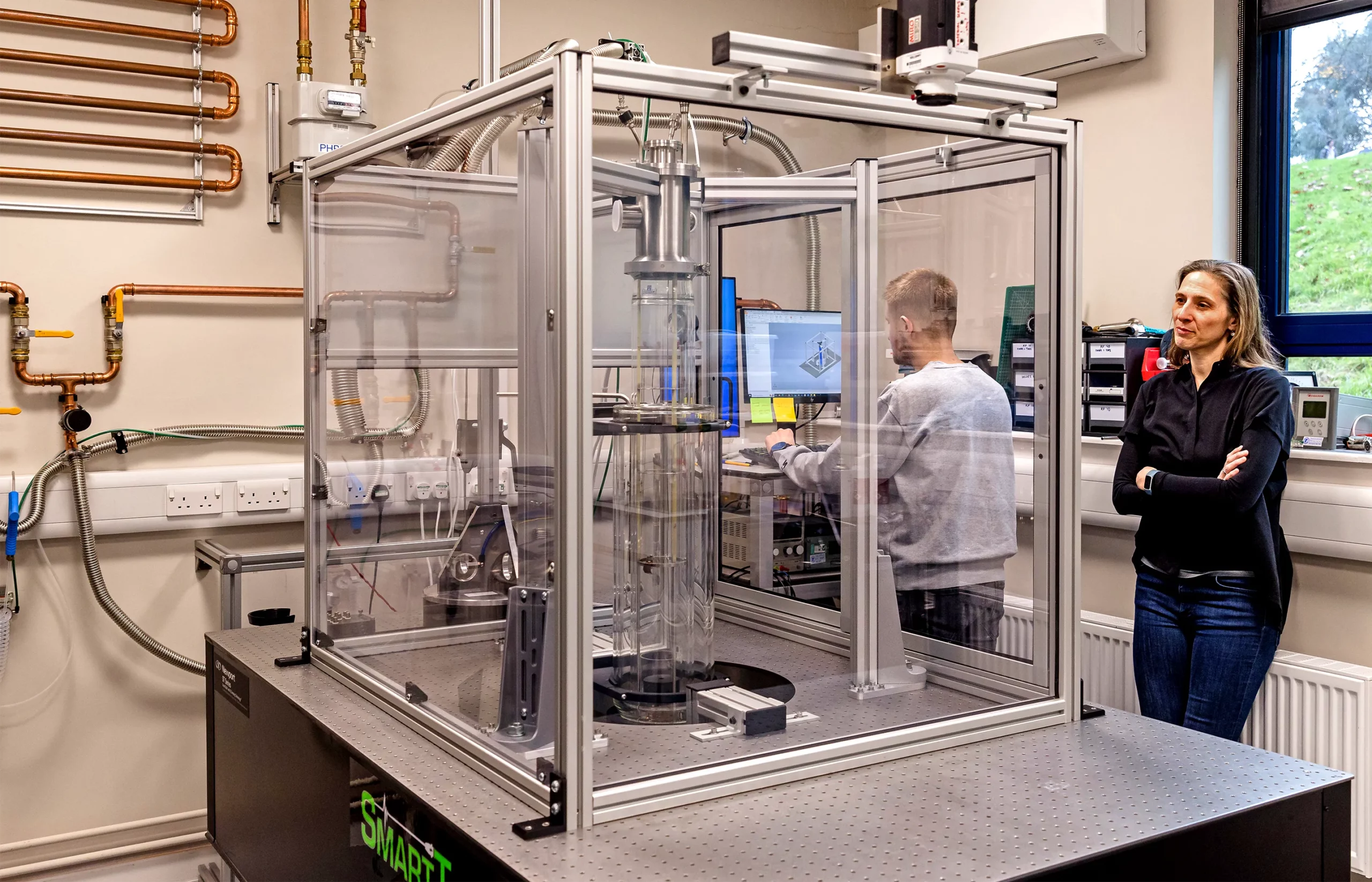 Weinfurtner stands next to a large glass box containing a superfluid helium tank that’s bolted to a vibration isolation table.