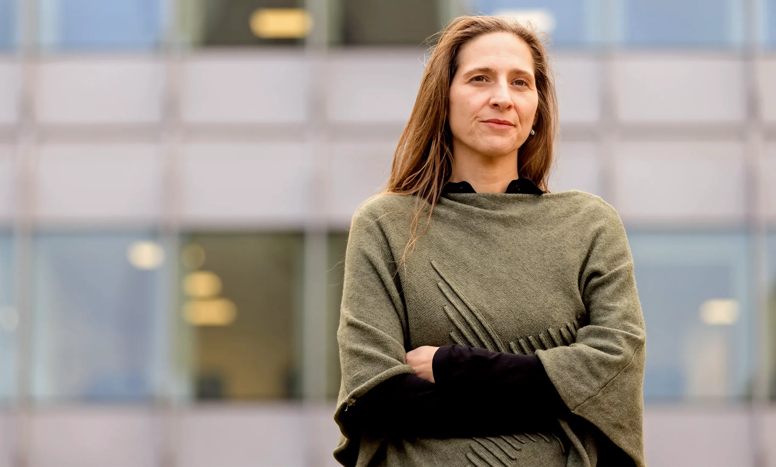 A portrait of a women with long brown hair and folded arms posing outside in front of a building.