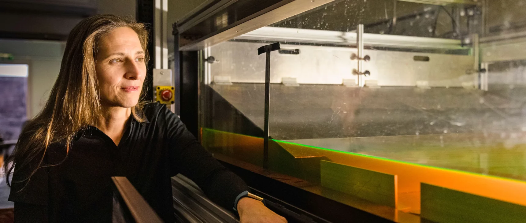 A woman stands in a darkened laboratory next to a shallow tank of water that glows green.