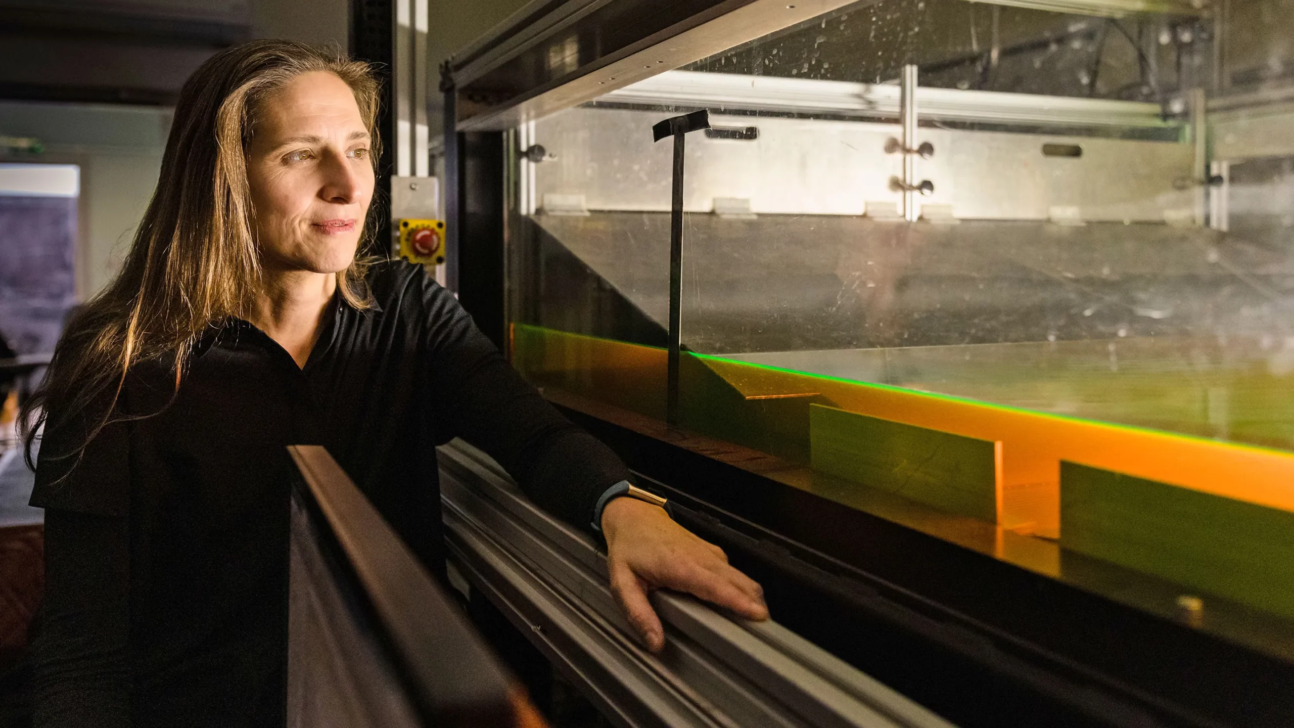 A woman stands in a darkened laboratory next to a shallow tank of water that glows green.