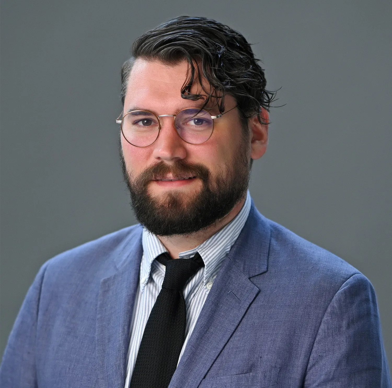 A bust portrait of a bearded young man wearing a black tie and blue suit jacket.