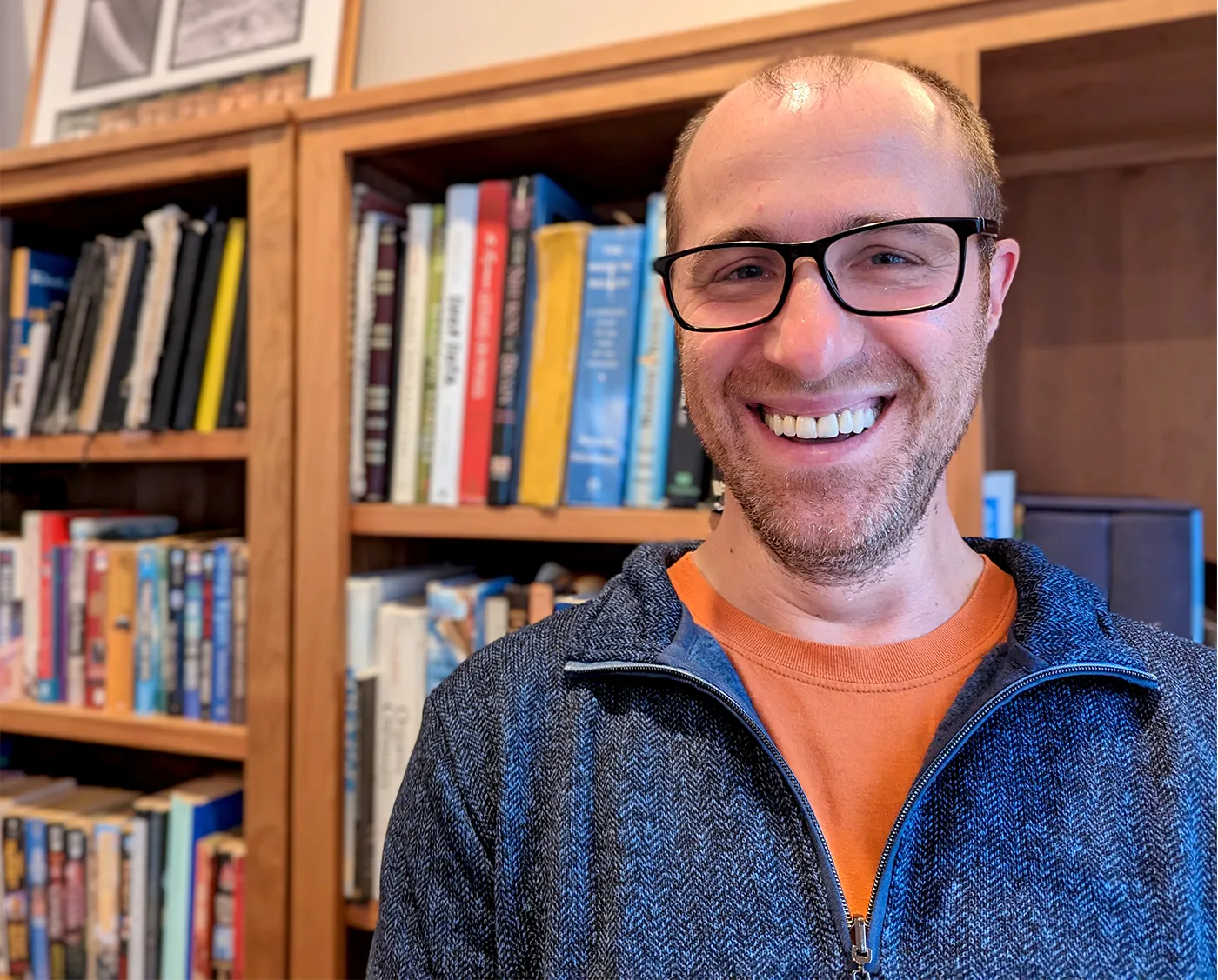 A photo of a man with light hair, a short beard, and dark-rimmed rectangular glasses smiling in front of a wooden bookshelf