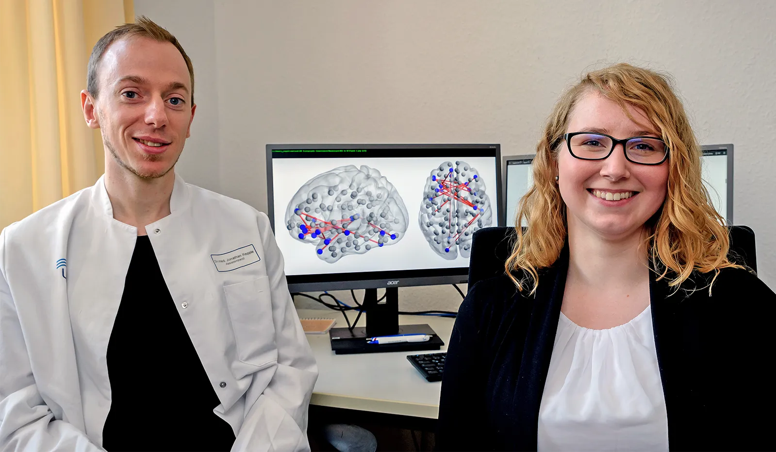 Jonathan Repple and Susanne Meinert of Goethe University sitting in front of a desk and a computer monitor displaying brain mapping data.