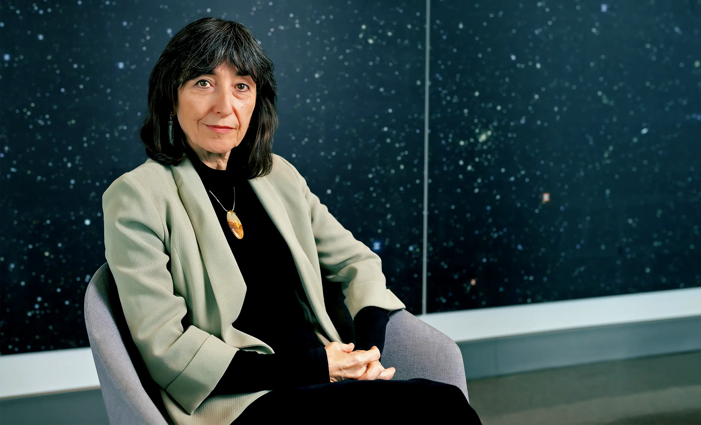 A woman with brown hair wearing a blazer sits in front of a blackboard.