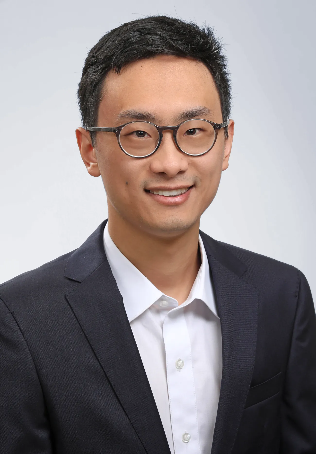 Photo of a young man in a white collared shirt, a dark dress jacket, and round glasses