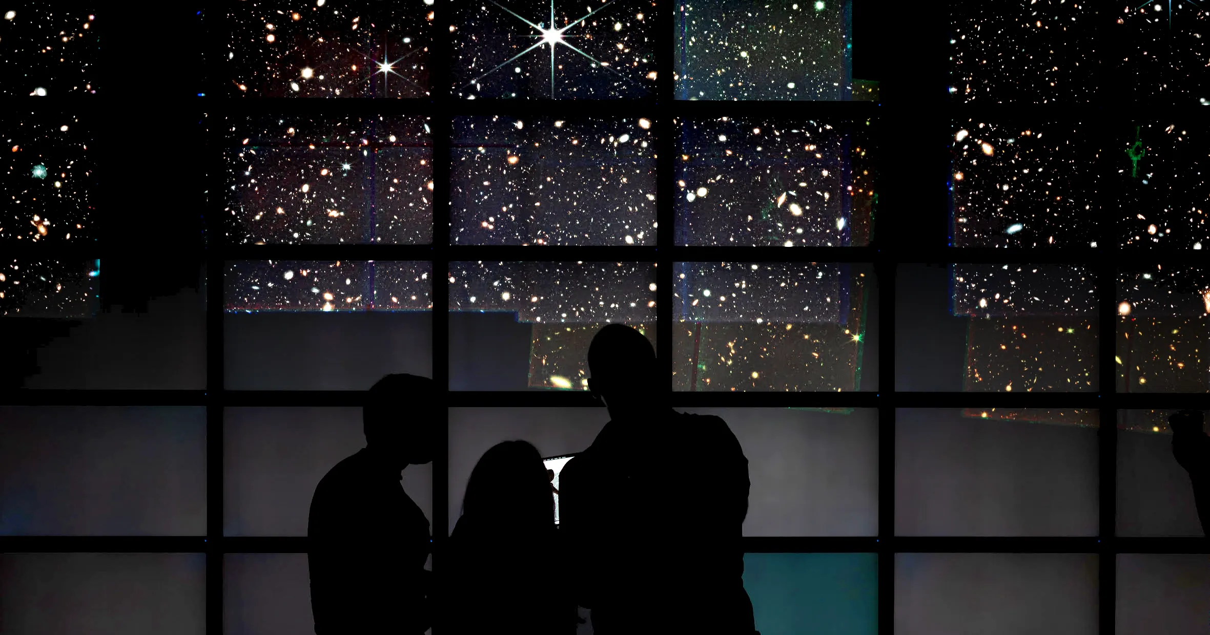 Shadows of scientists are seen in front of a panel of screens displaying images of the universe.]