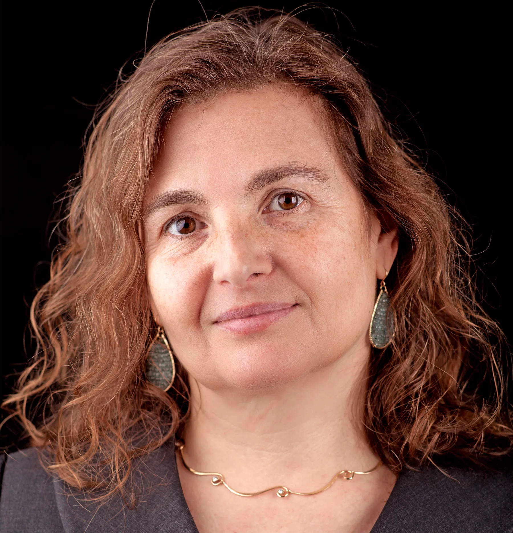 a woman with short, curly reddish brown hair with gold and green teardrop earrings