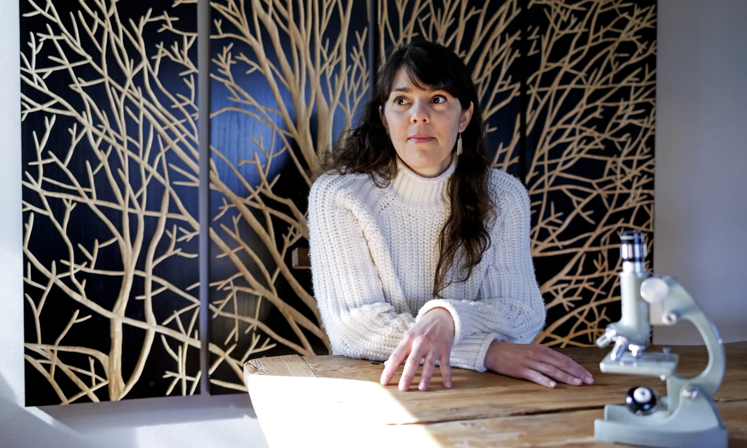 Red Horse seated at a table with a microscope in front of her and artwork of a tree behind her.