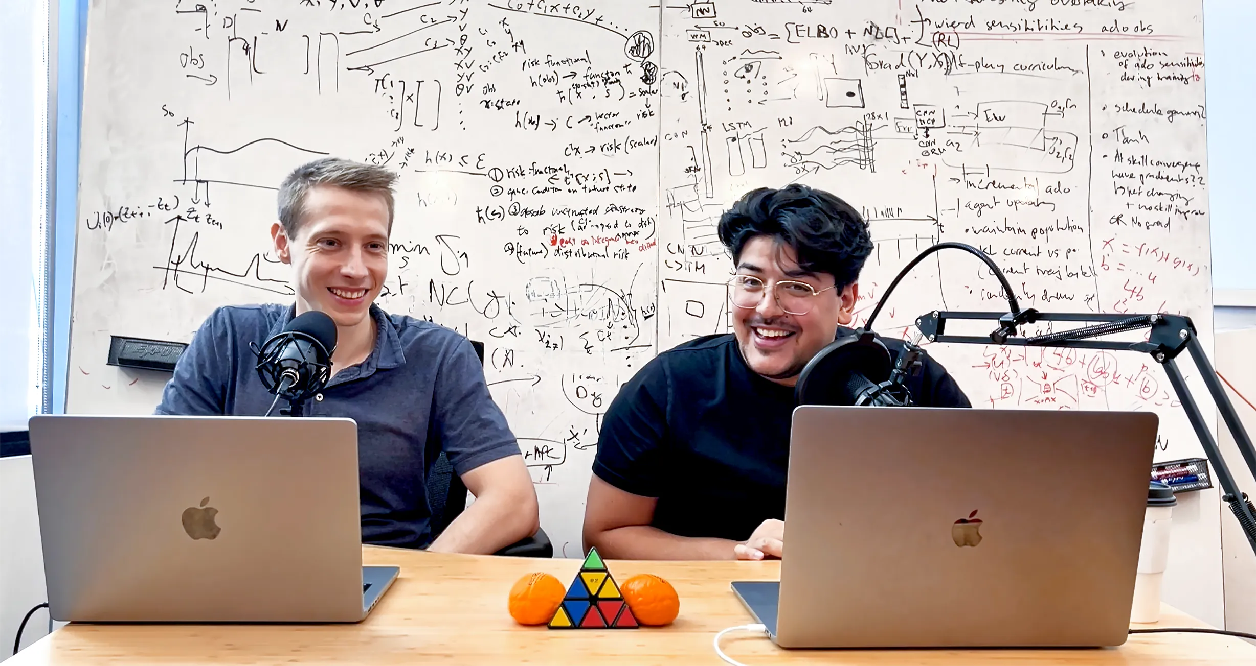 two men sit in front of a whiteboard covered in equations, and behind microphones and MacBook computers