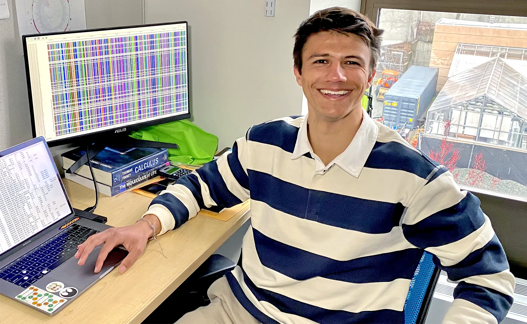 Gozashti sitting at his office desk in front of monitors showing his genomic work.