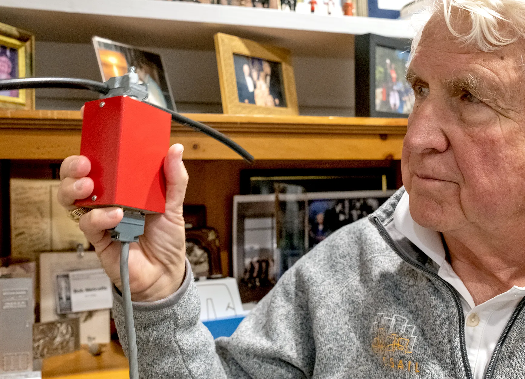 Bob Metcalfe holds a red box with wires coming out of it, in front of a shelf of photos.