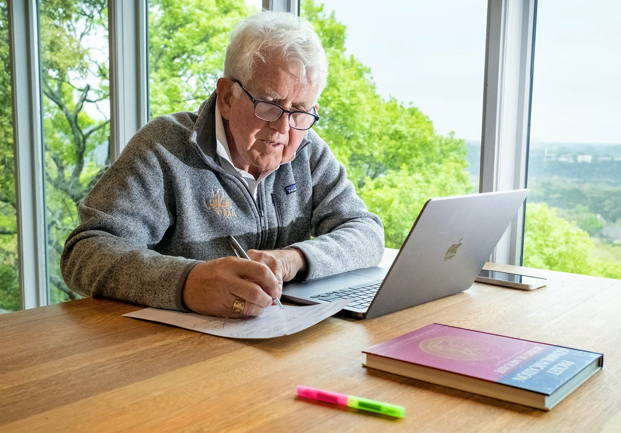 Bob Metcalfe sits at a wooden table using a laptop with trees in the background.