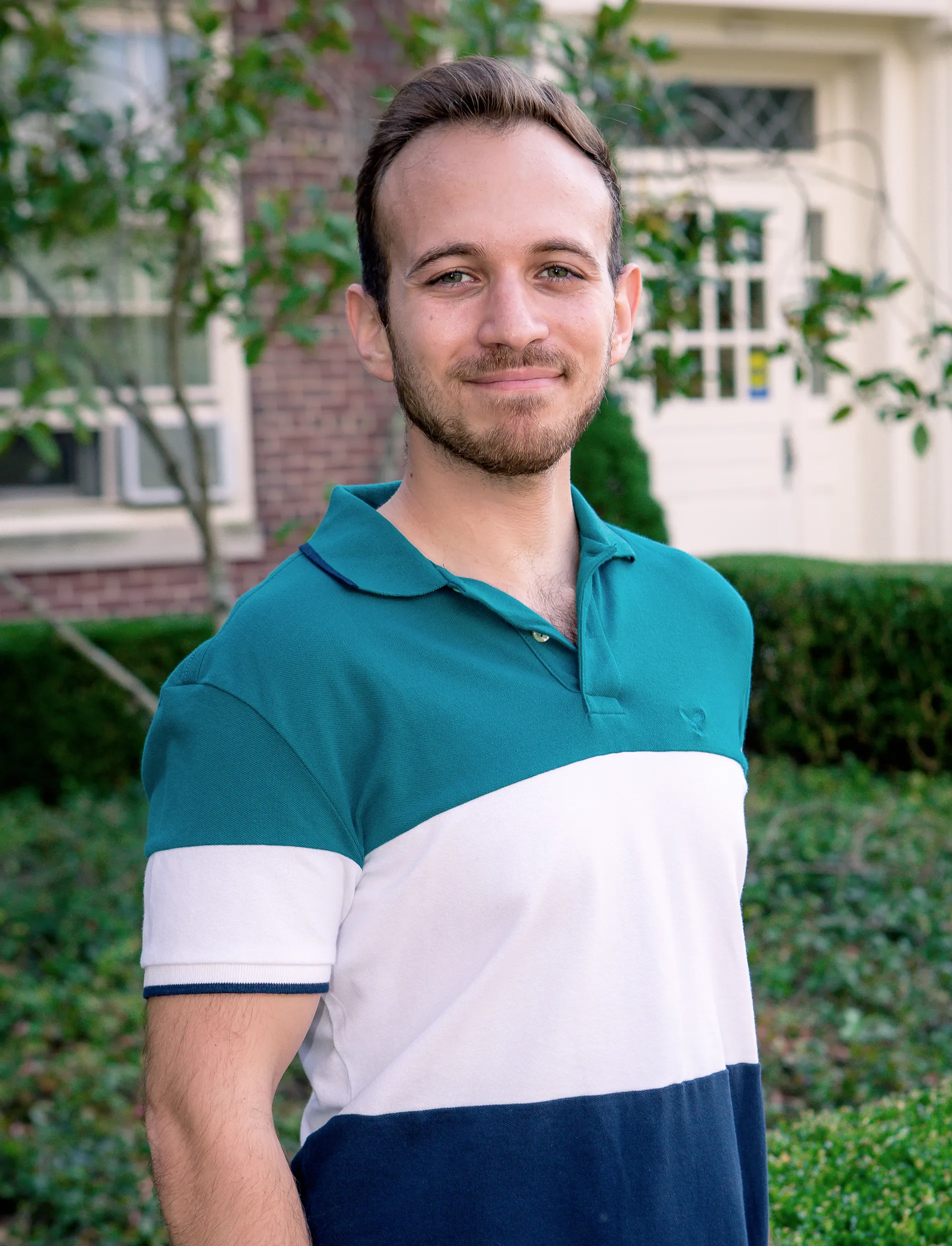 a man in a striped polo and short beard in front of a brick building and bushes