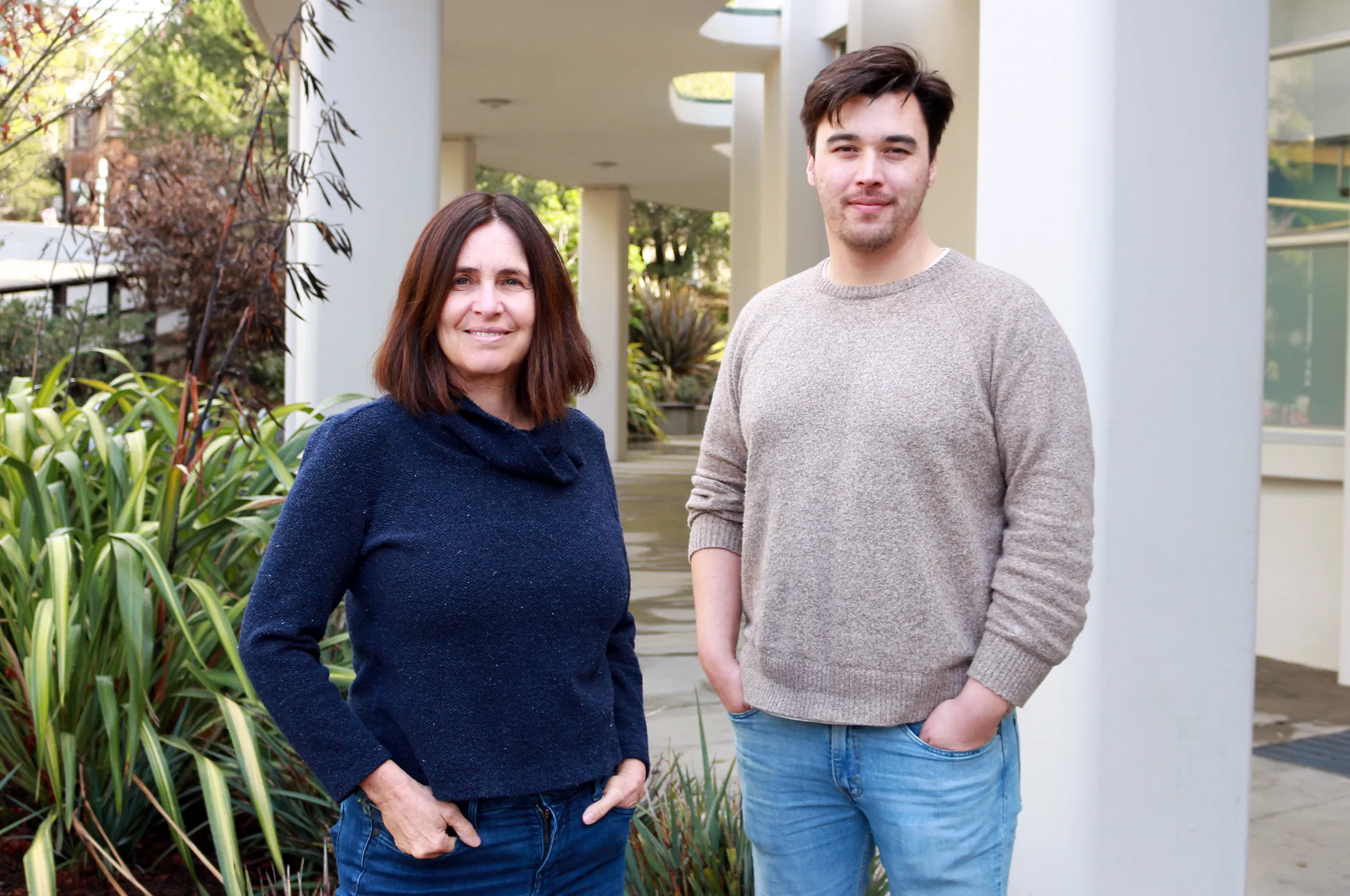 a woman with straight brown hair and a young man with a brown sweater and short beard smiling in front of a university building