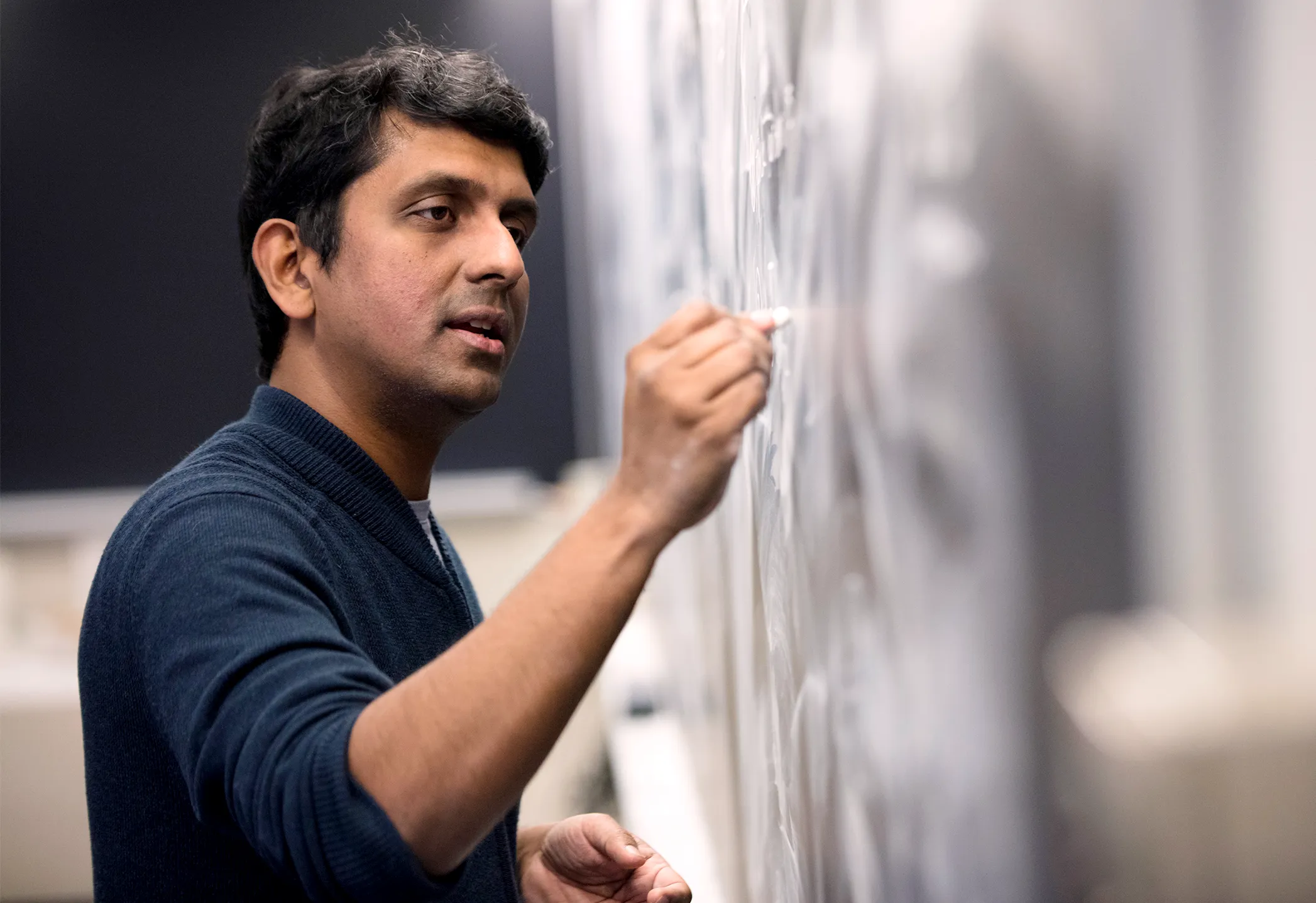 a medium-skinned man with dark hair and a blue sweater writes on a chalkboard