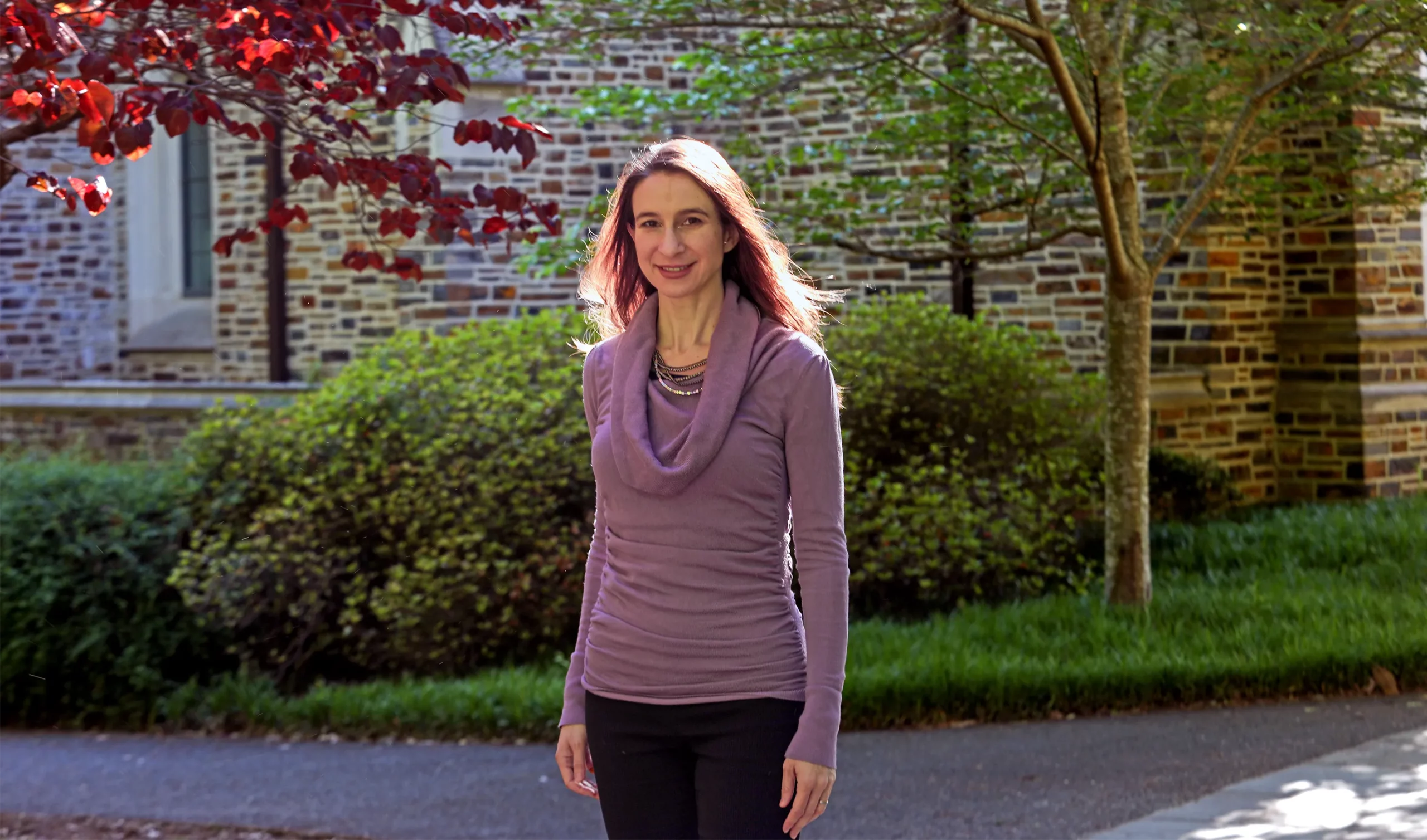 Cynthia Rudin, in a purple sweater, stands outdoors in front of a stone wall.