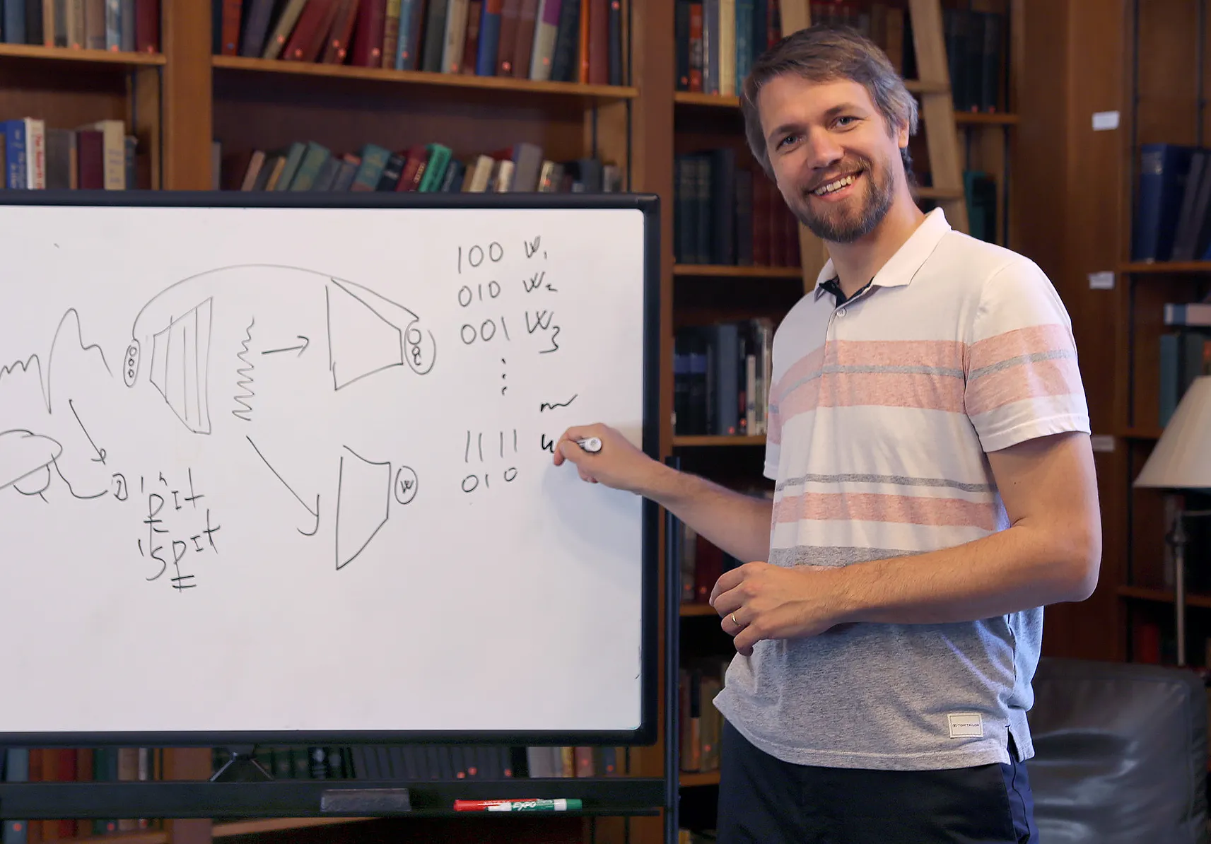 Gašper Beguš in a striped polo shirt with a whiteboard in front of books.