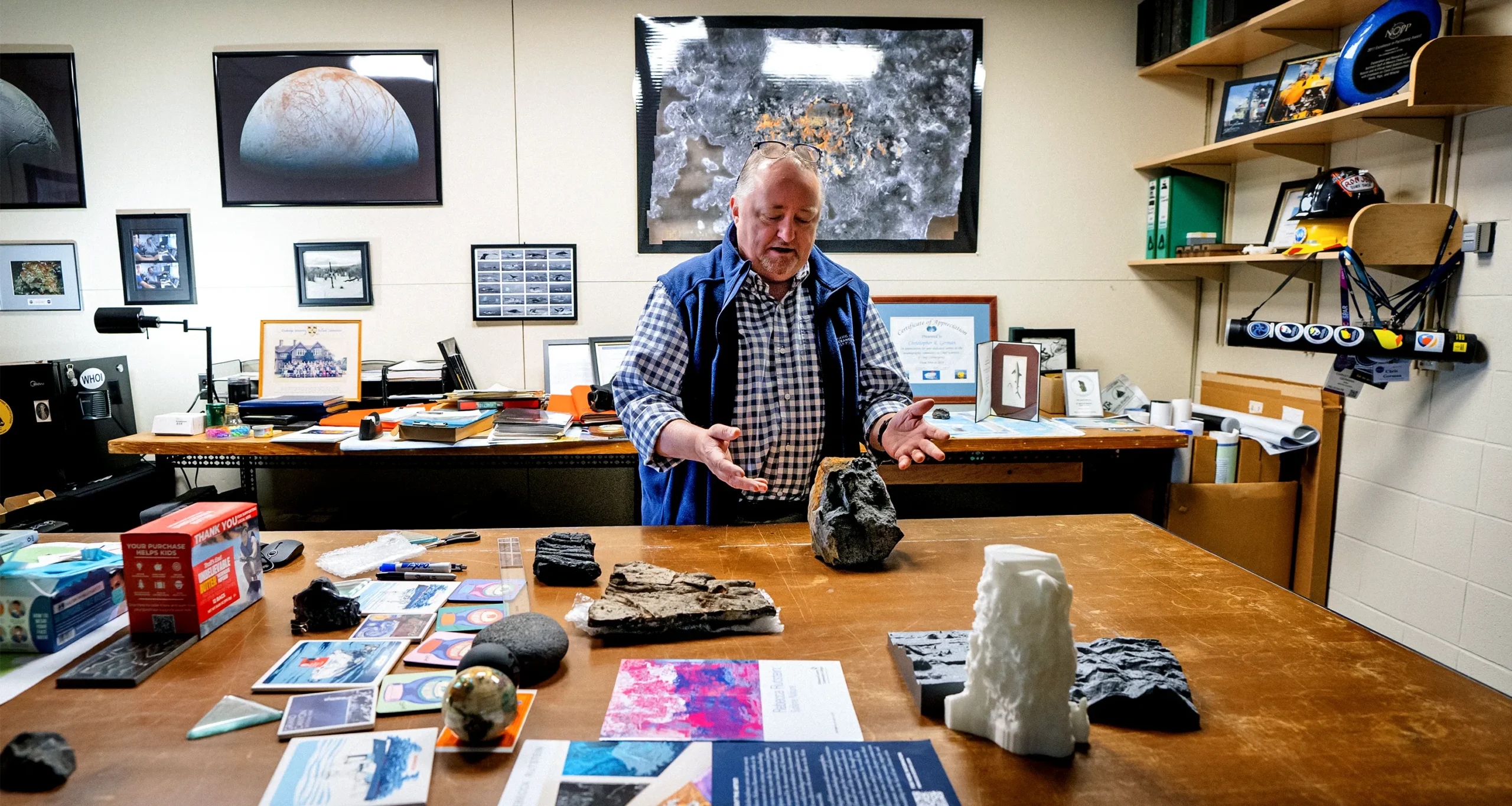 Chris German gesturing with a large sample of rock recovered from the seafloor while standing beside a large table in his office that is covered with samples and images and models of the seafloor.