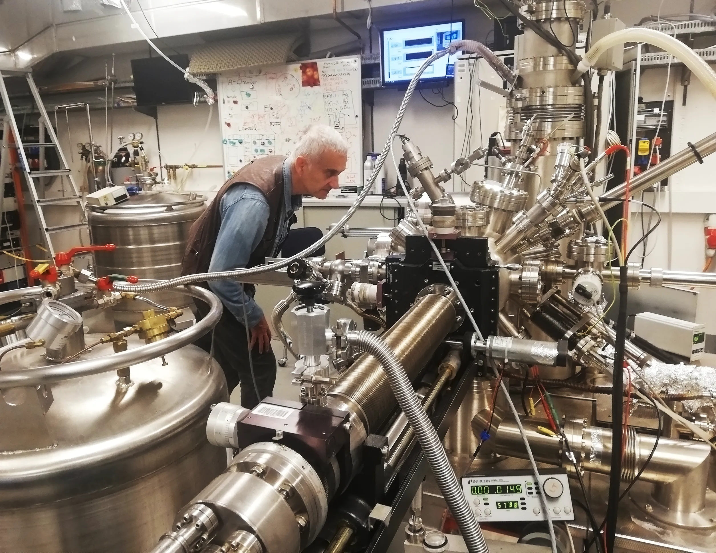 An image of Gábor Domokos, staring at a scanning tunneling microscope. The instrument is big, shiny tangle of tubes. He's in a room filled with tanks, a ladder, and a whiteboard.