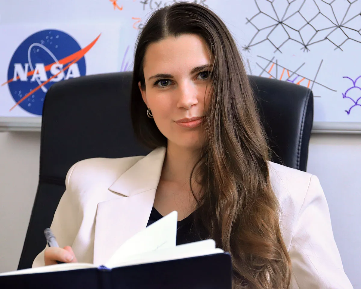 A portrait of Krisztina Regős. She is in front of a white board filled with sketched patterns and is wearing a white jacket and smiling. The NASA logo is visible over her shoulder.