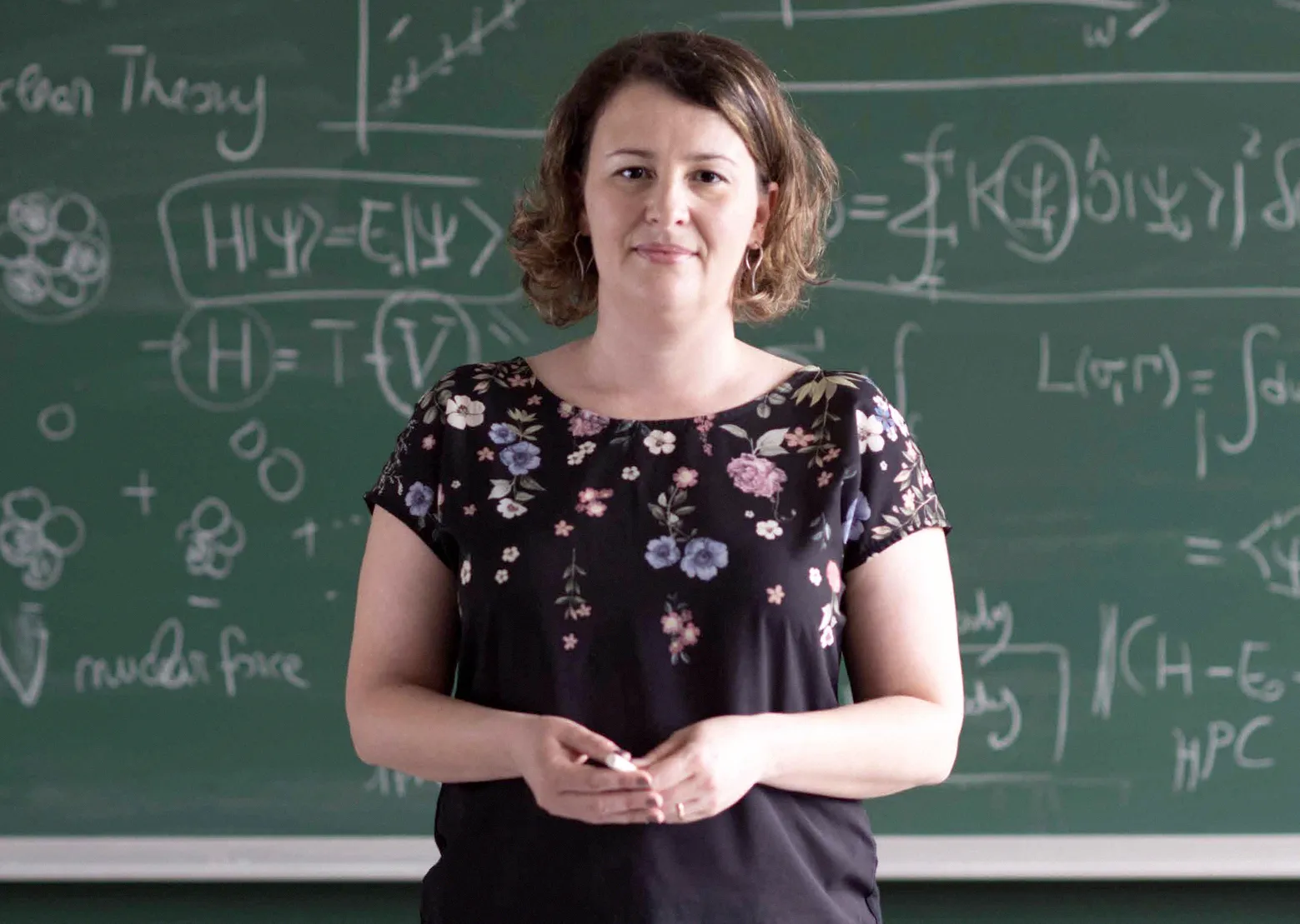 Sonia Bacca, wearing a black, flowered shirt, stands in front of a chalkboard filled with equations. She's holding a piece of chalk.