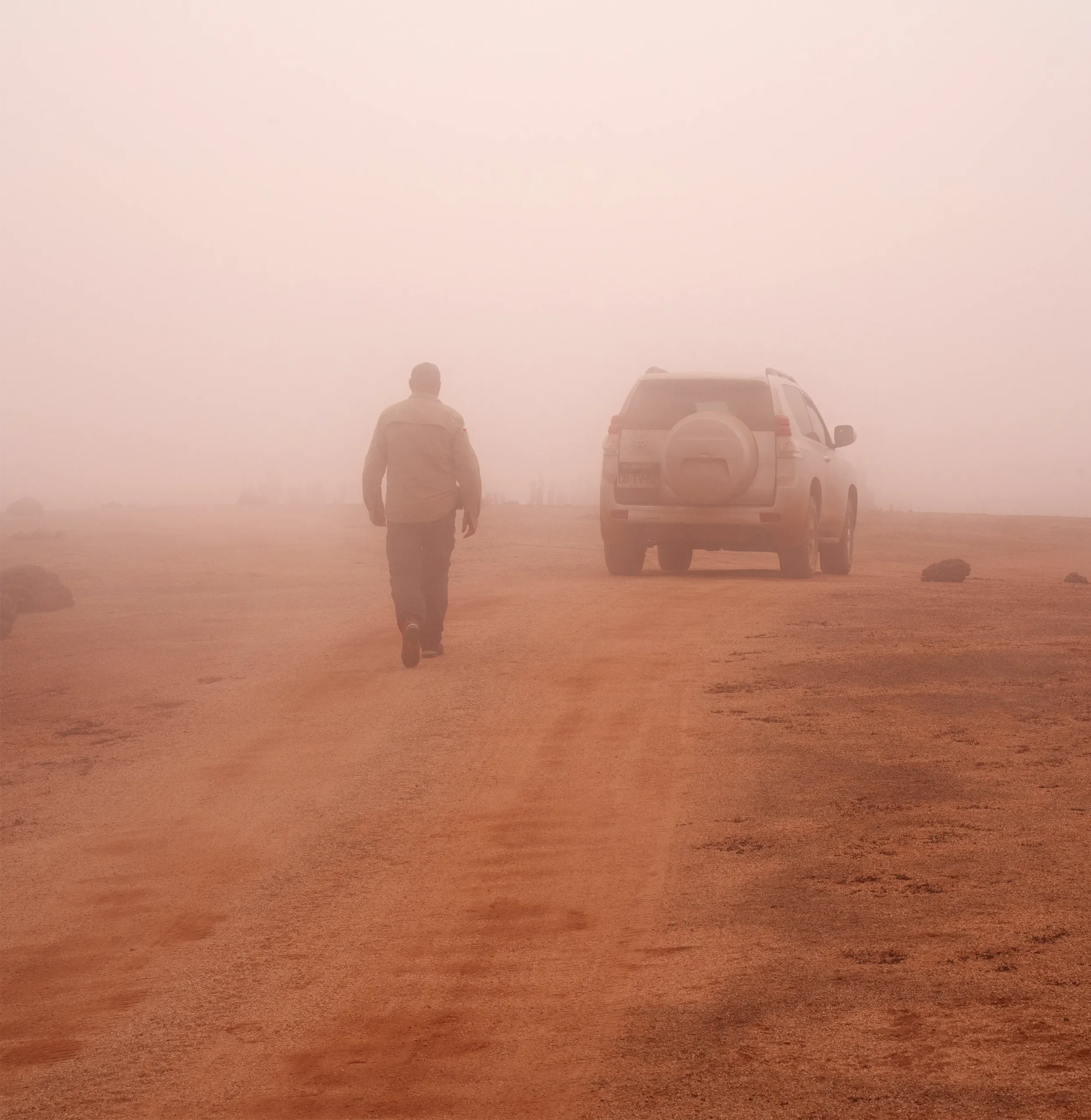 Male human figure walking away from the viewer and toward a car through thick fog. Footprints in the desert crust are visible behind him.