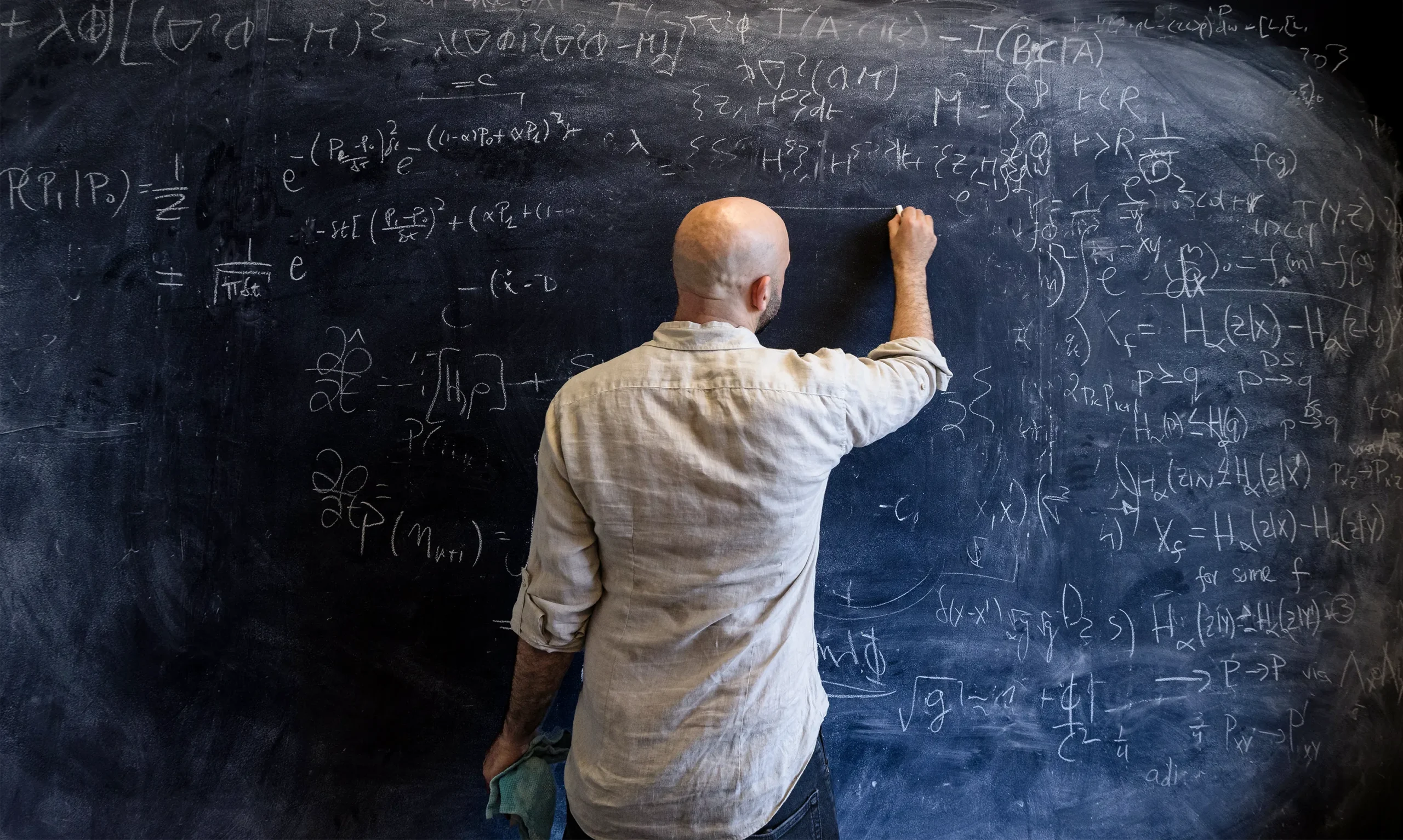 Oppenheim writing on a blackboard that is stuffed with equations. His back is to the camera.