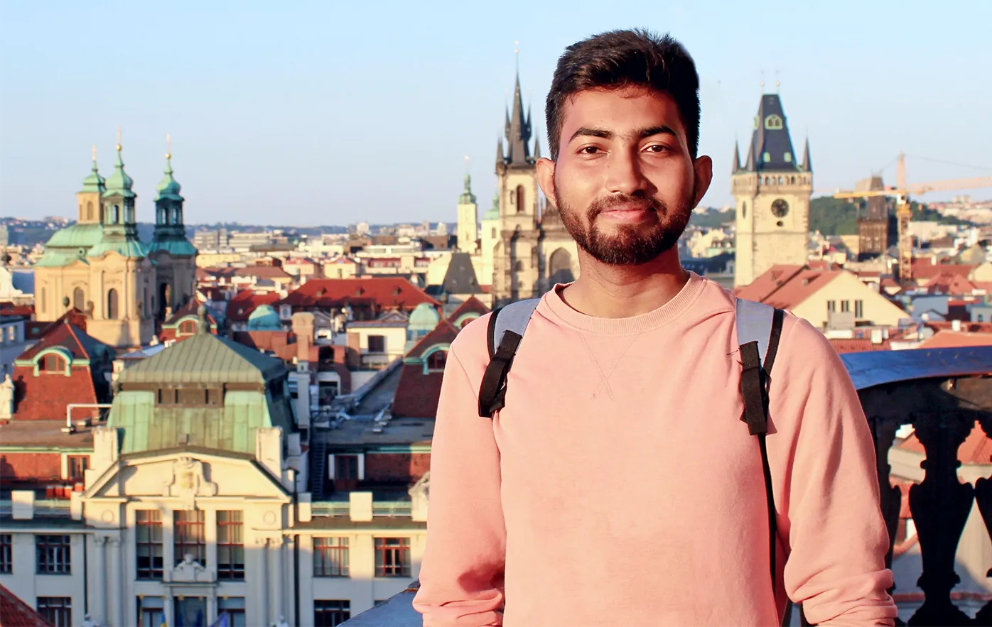 Biswas is outside on a balcony, with a city skyline in the background. He is wearing a backpack and a peach sweatshirt.