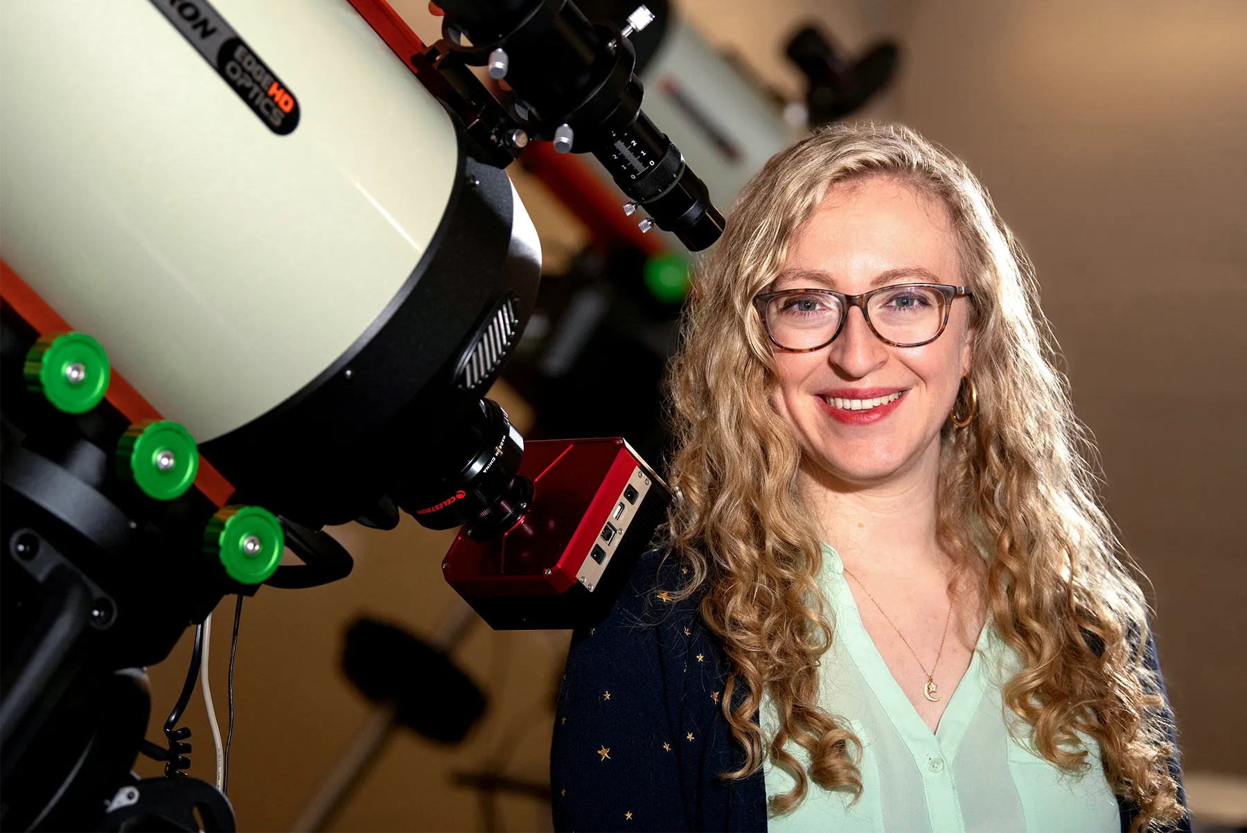 Villadsen is standing near the base of a telescope, smiling. She’s wearing a mint green shirt. The telescope is angled up and to the left.