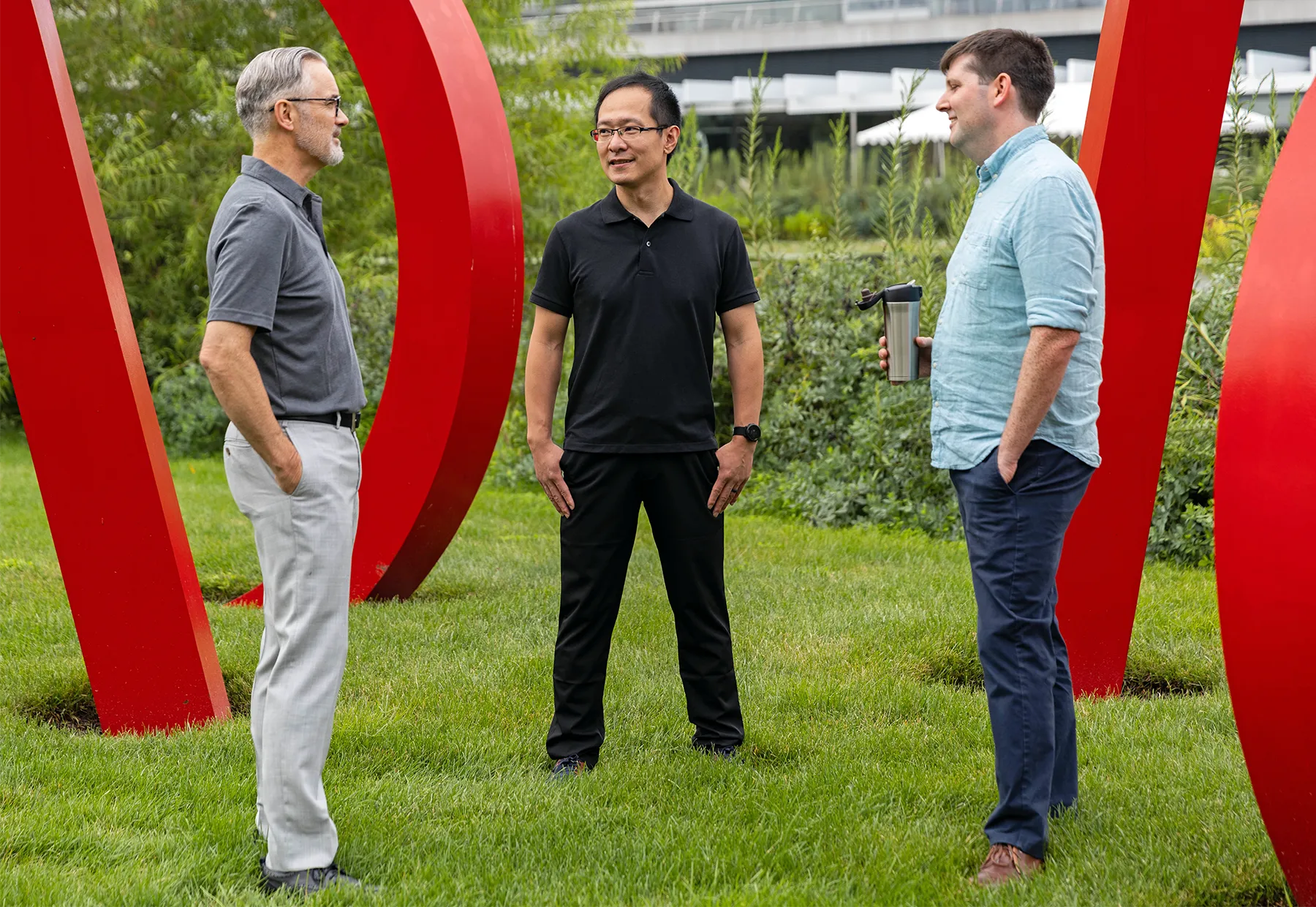 Three scientists stand facing one another on a grassy campus, surrounded by red modern art sculpture.