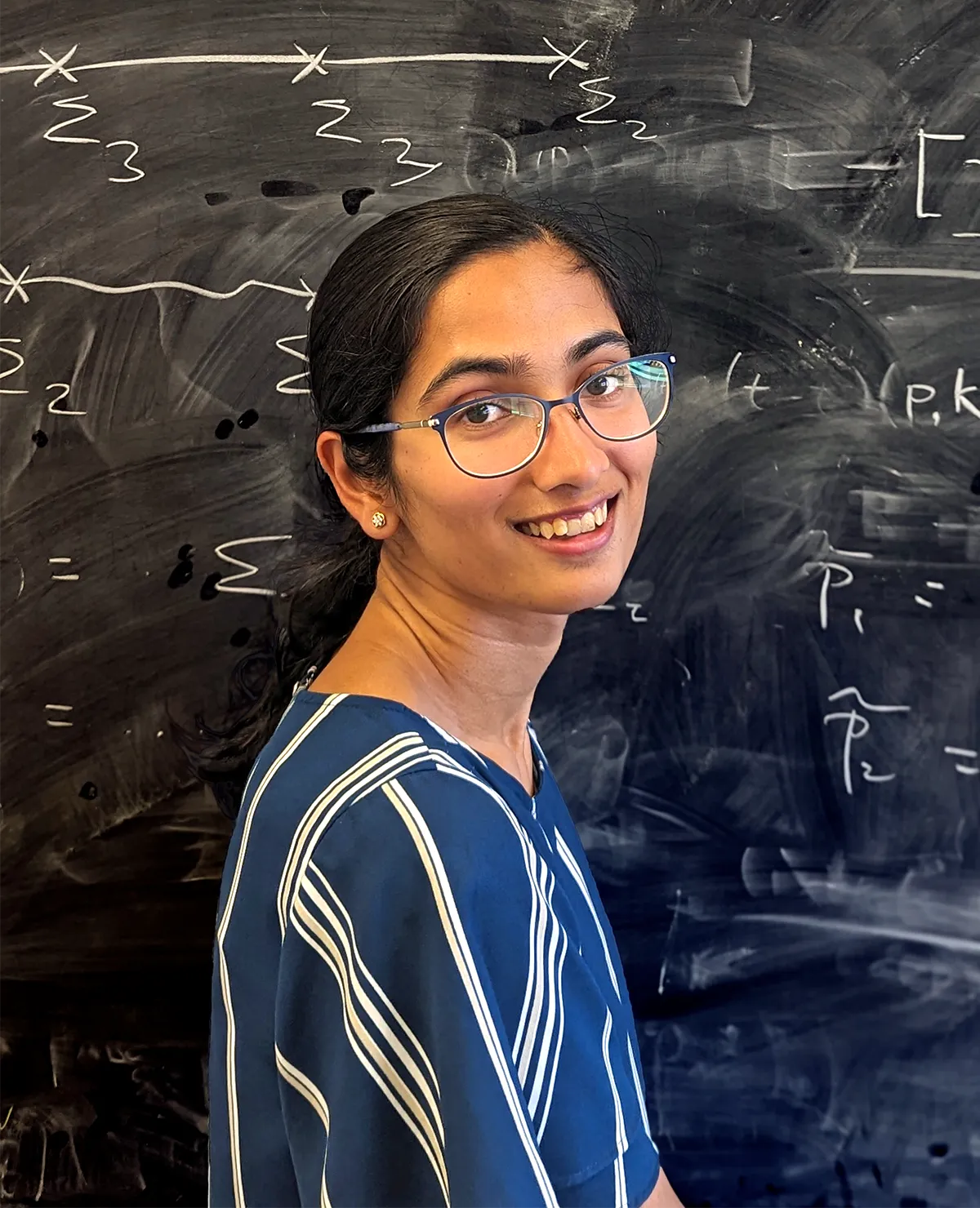 A woman stands in front of a chalkboard filled with equations.