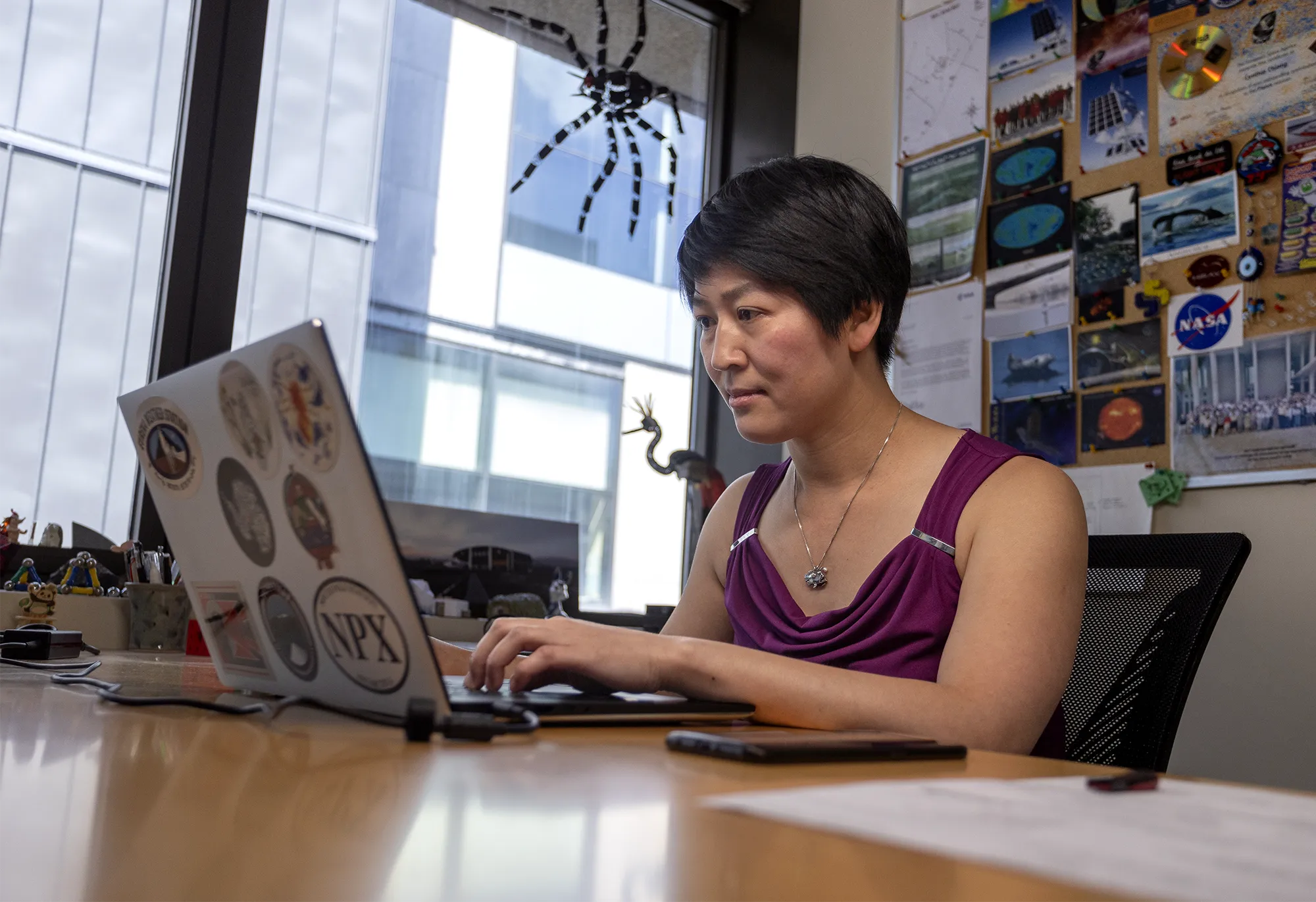 Chiang is at her desk, working on a laptop that’s covered in stickers. Behind her is a board filled with postcards, photos and mementos from various projects, including a prominent NASA sticker and images of Antarctic wildlife. A large toy spider hangs in the window.