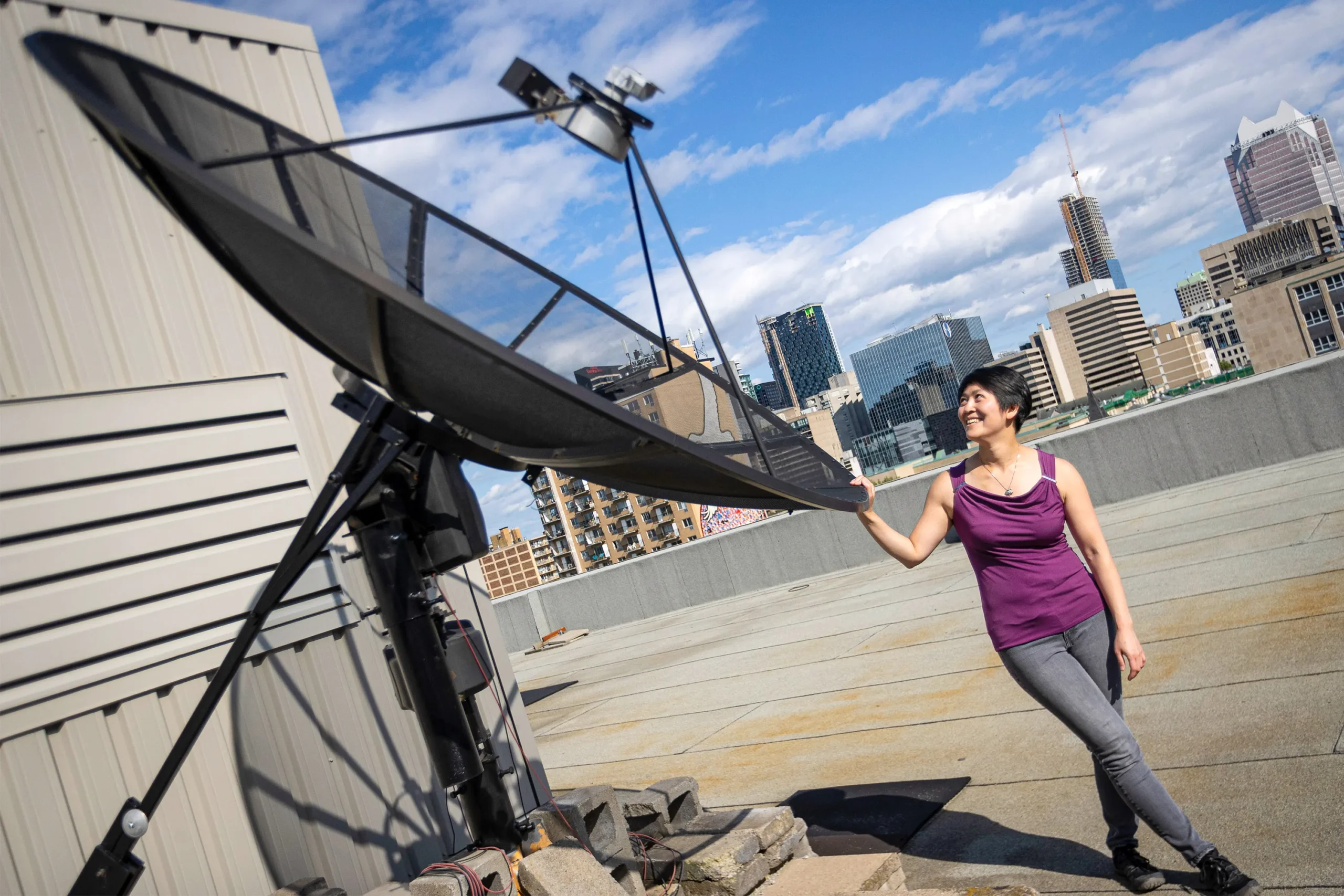 Chiang is on the roof of McGill’s Rutherford building, standing next to a parabolic antenna that is taller than she is. She is looking up at the antenna and smiling. Montreal is in the background, along with a partly cloudy blue sky.