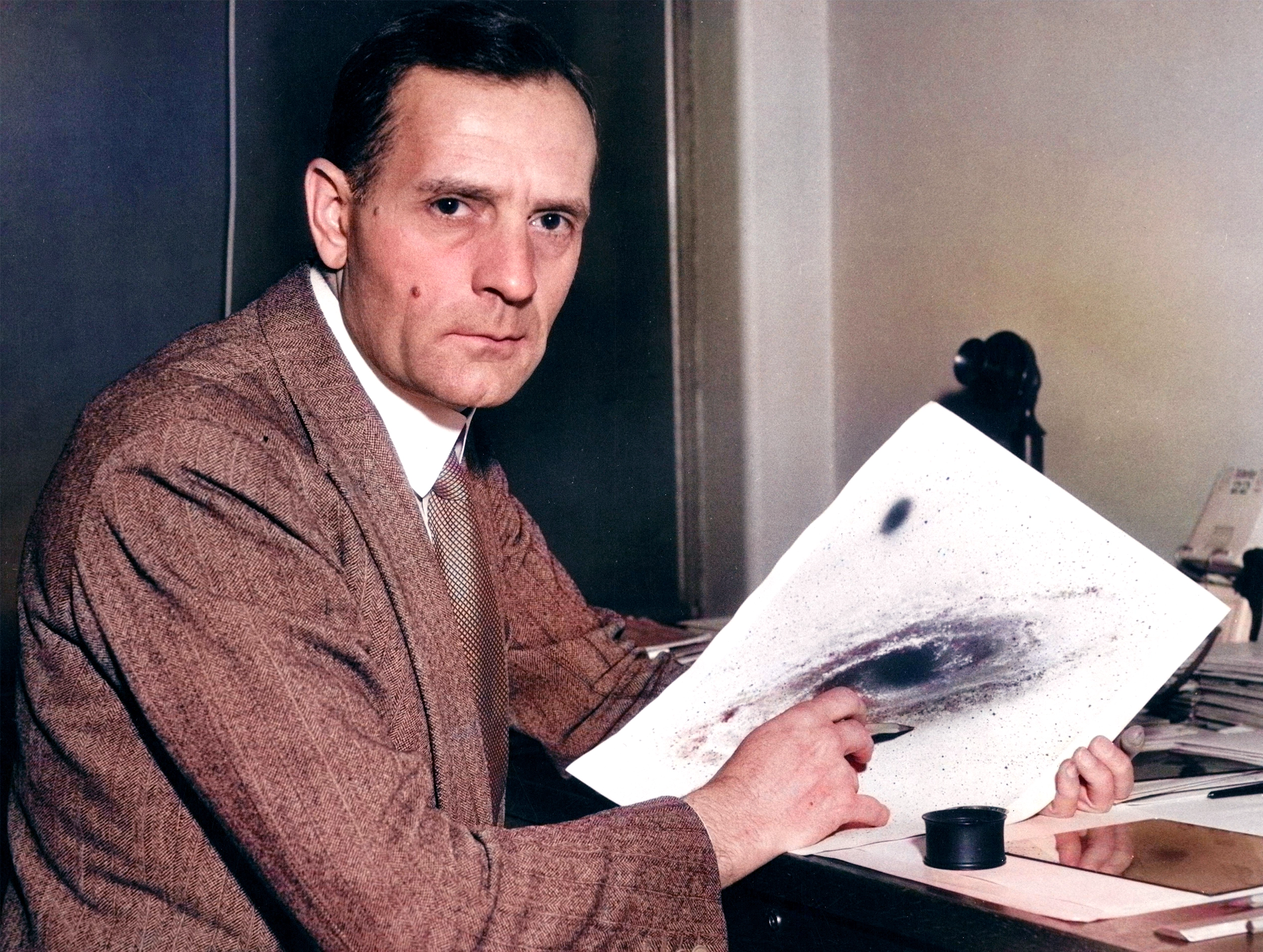 Edwin Hubble sits at his desk. He’s looking at the camera and pointing to a black-and-white image of a galaxy.