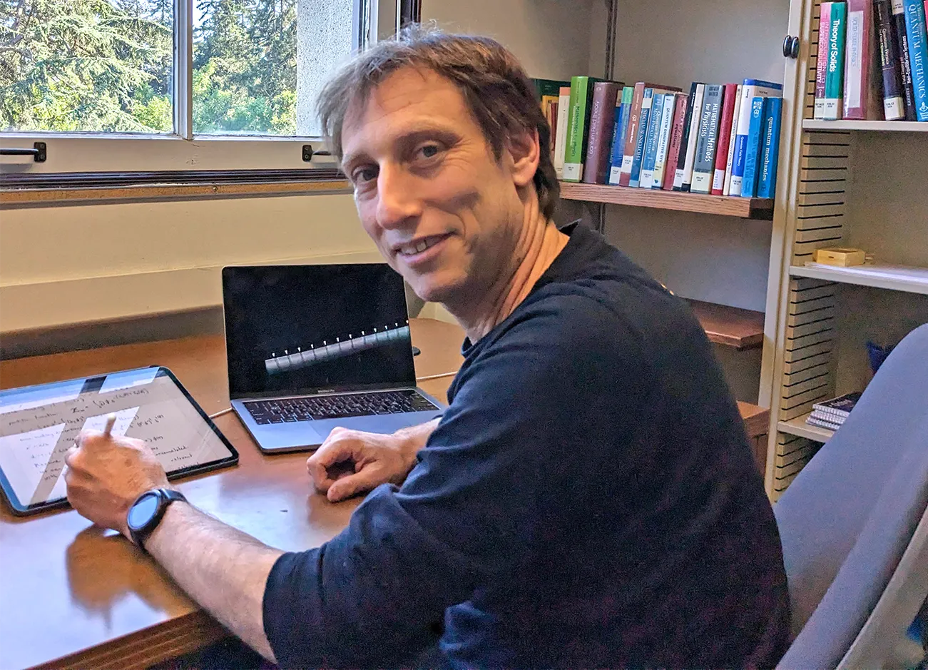 Ehud Altman sitting at his desk. He has swiveled partway around to smile at the camera. In the background are a bookshelf and a window, through which evergreen trees are visible.