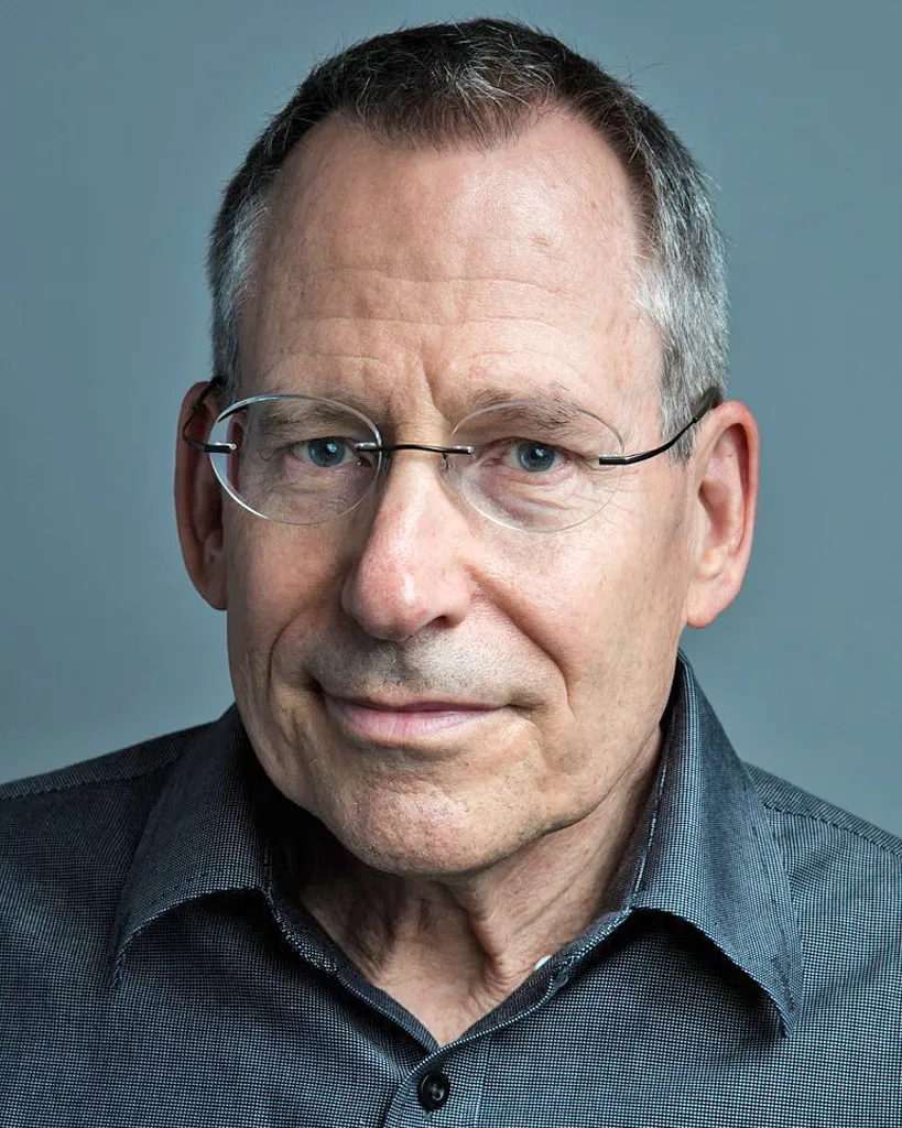 John Preskill in a gray shirt, posing in front of a blue background and smiling.