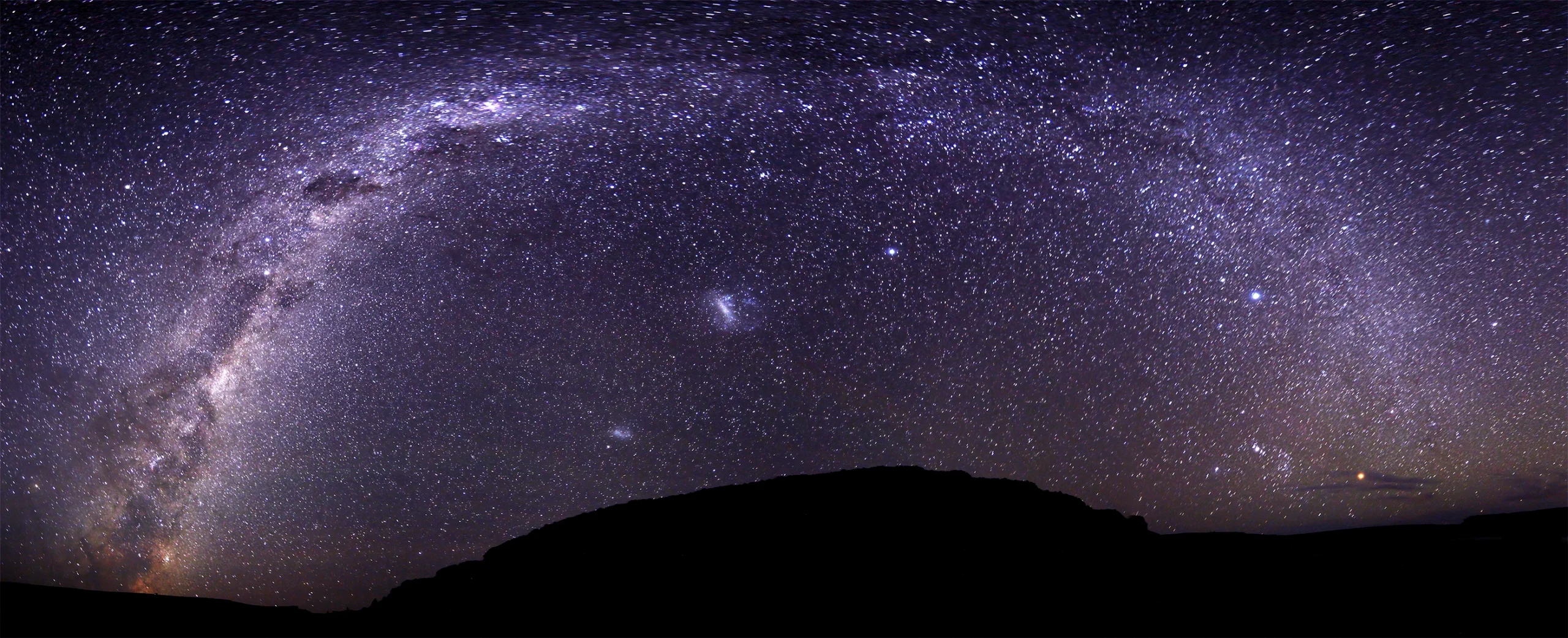 A panoramic image of the Milky Way at night, taken from Argentina. A small hill is in the foreground, in silhouette. The galaxy arches across the sky. Beneath it are two prominent smudges: the Large and Small Magellanic clouds.